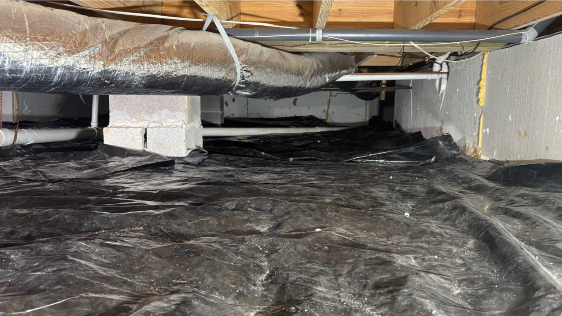 Crawl space with black plastic vapor barrier, exposed pipes, and a white support pier under the floor