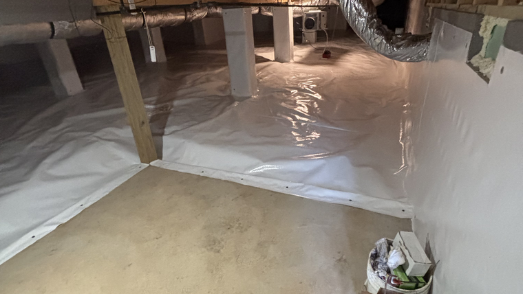 Crawl space with plastic sheeting on the floor, exposed beams, and a bucket near the wall.