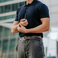 Man in a black polo shirt adjusting his gray trousers outdoors in front of a modern building