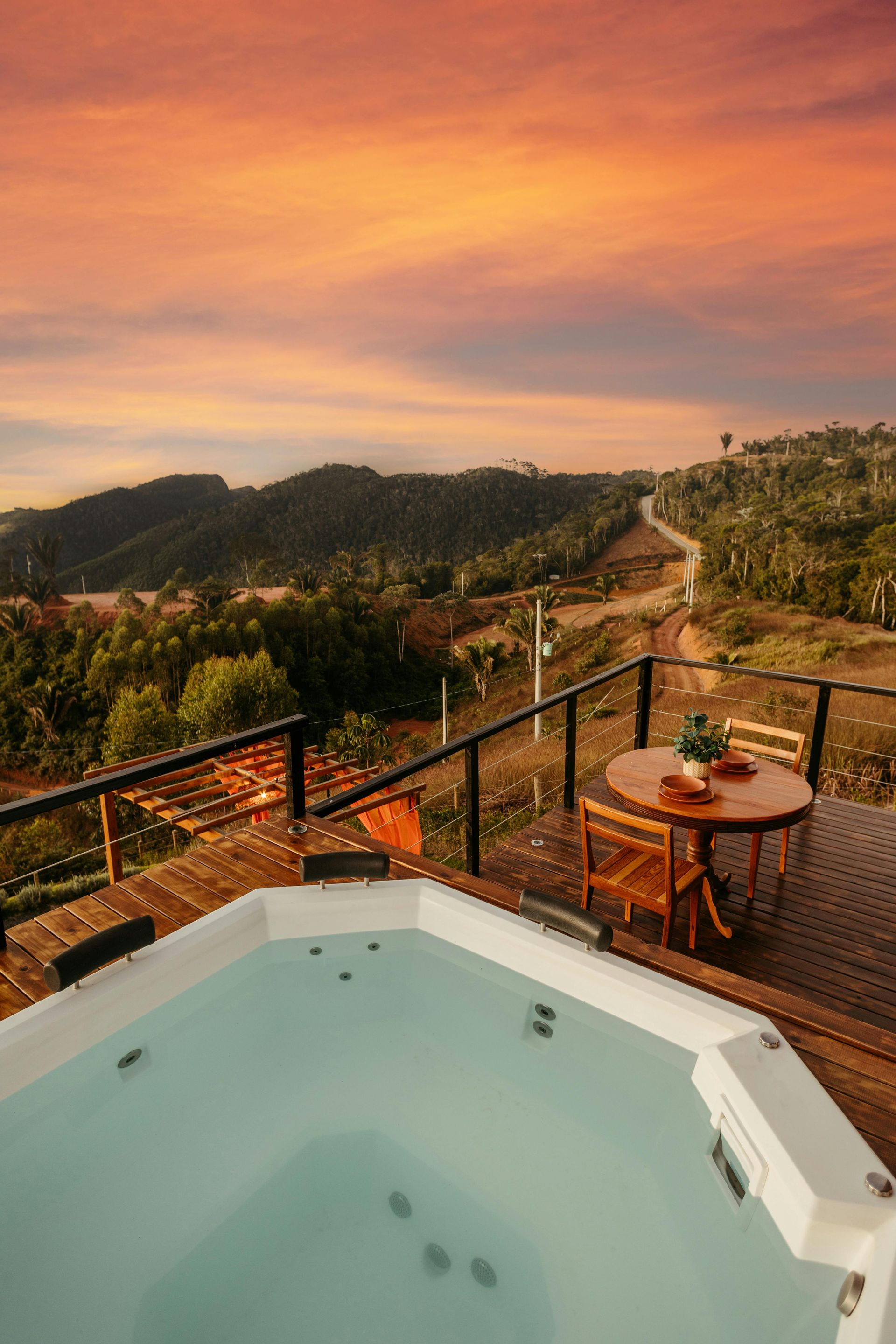 Hot tub on a deck overlooking a hilly landscape at sunset, with a small table and chairs nearby.