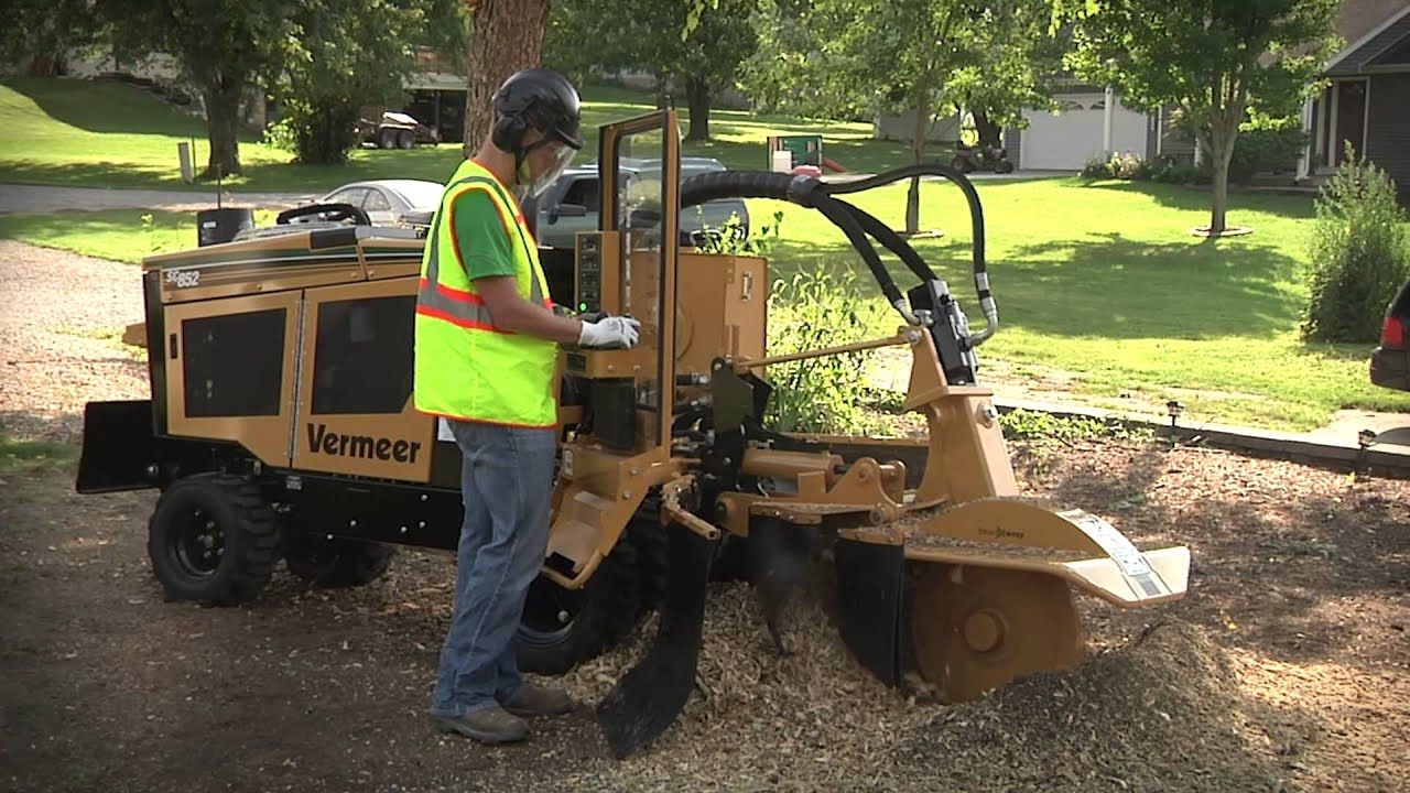 Man operating a stump grinder on a paved surface. He wears a safety vest, and the machine is beige and black.