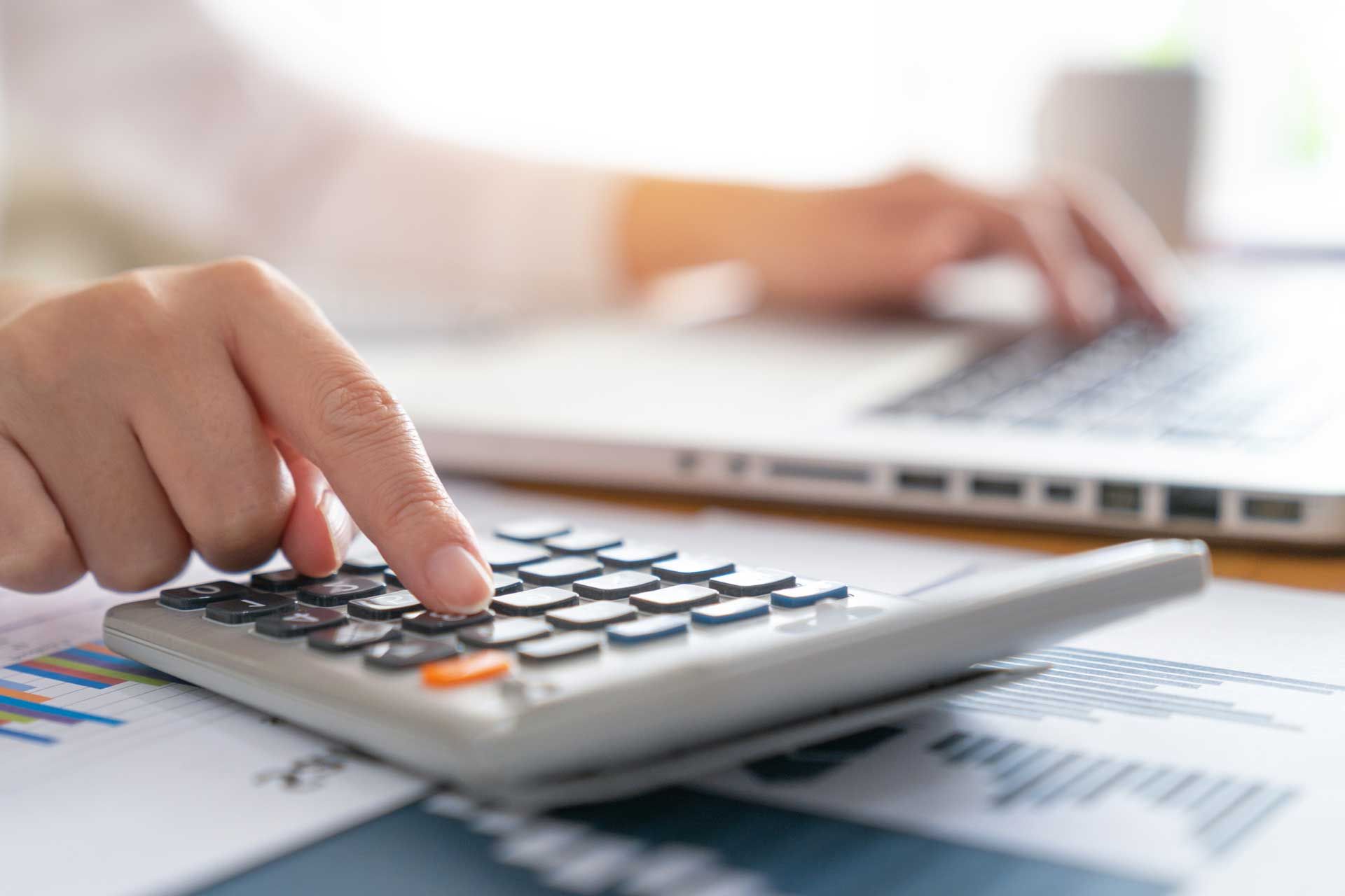 Person using a calculator with a laptop in the background, documents on the desk.