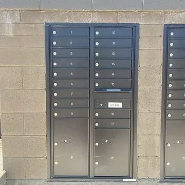 A dark metal wall-mounted mailbox unit with numbered compartments, set against a concrete block wall.