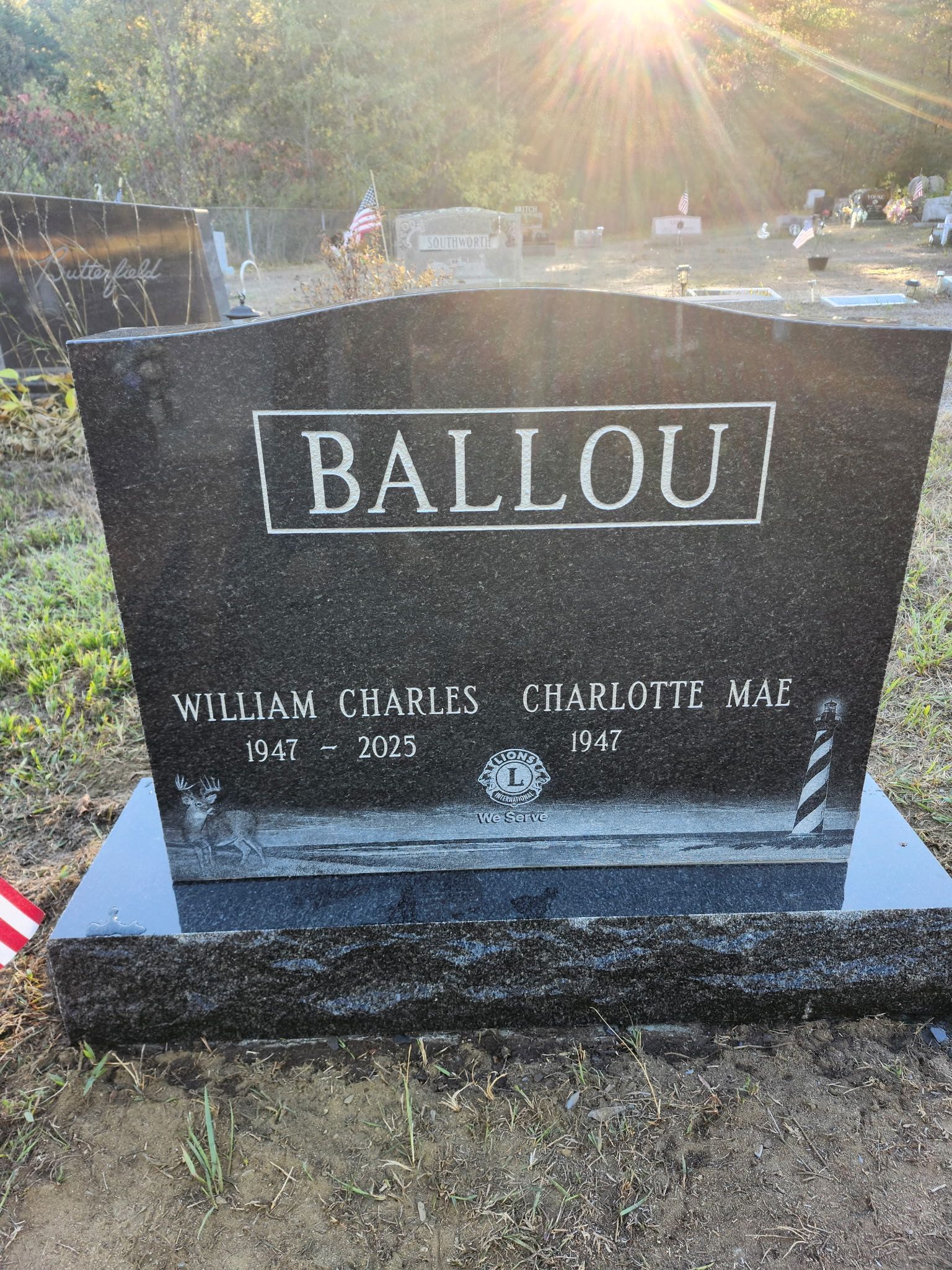 Black granite gravestone engraved “BALLOU” with flowers and sunlit cemetery background