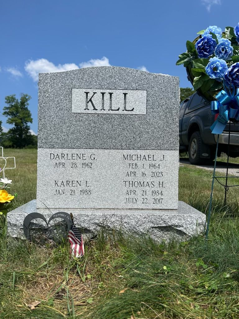Gravestone with “KILL” engraved, decorated with flowers, on a sunny roadside memorial.