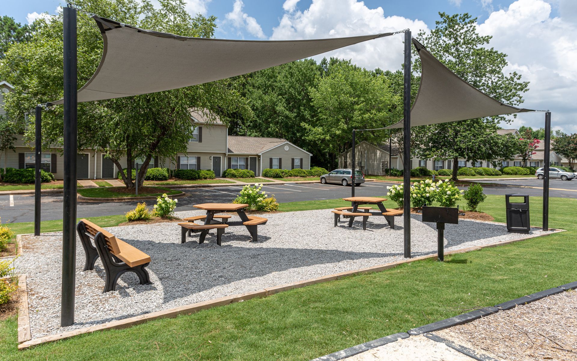 An outdoor recreation area with picnic tables and a park bench under two grey shade sails on a gravel base.