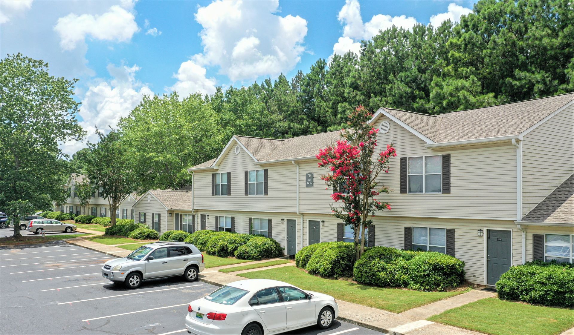 A row of two-story light-colored apartment buildings with parking spaces, trees, and blue sky in the background.