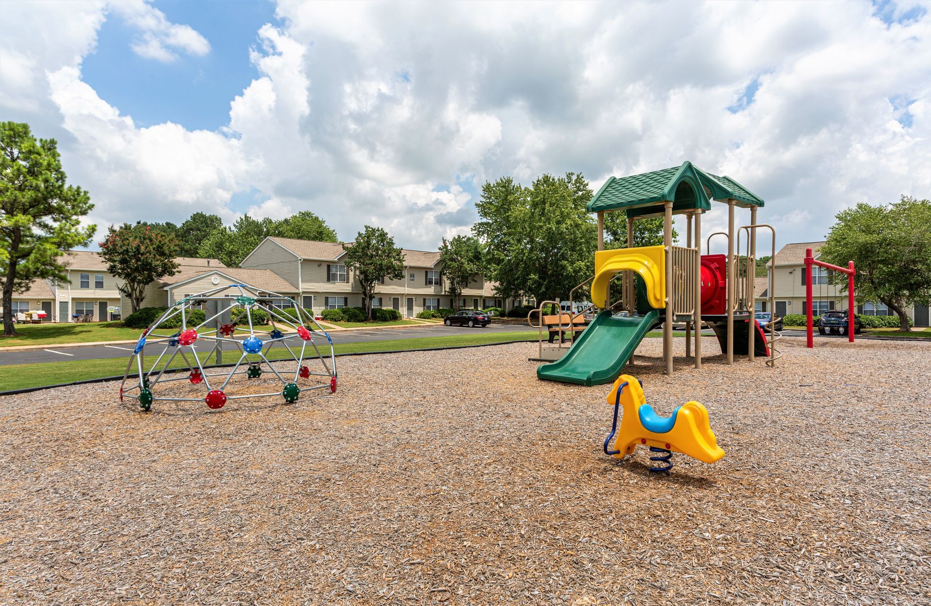 An outdoor playground with a climbing dome, slide structure, and yellow spring rider on a wood chip base.