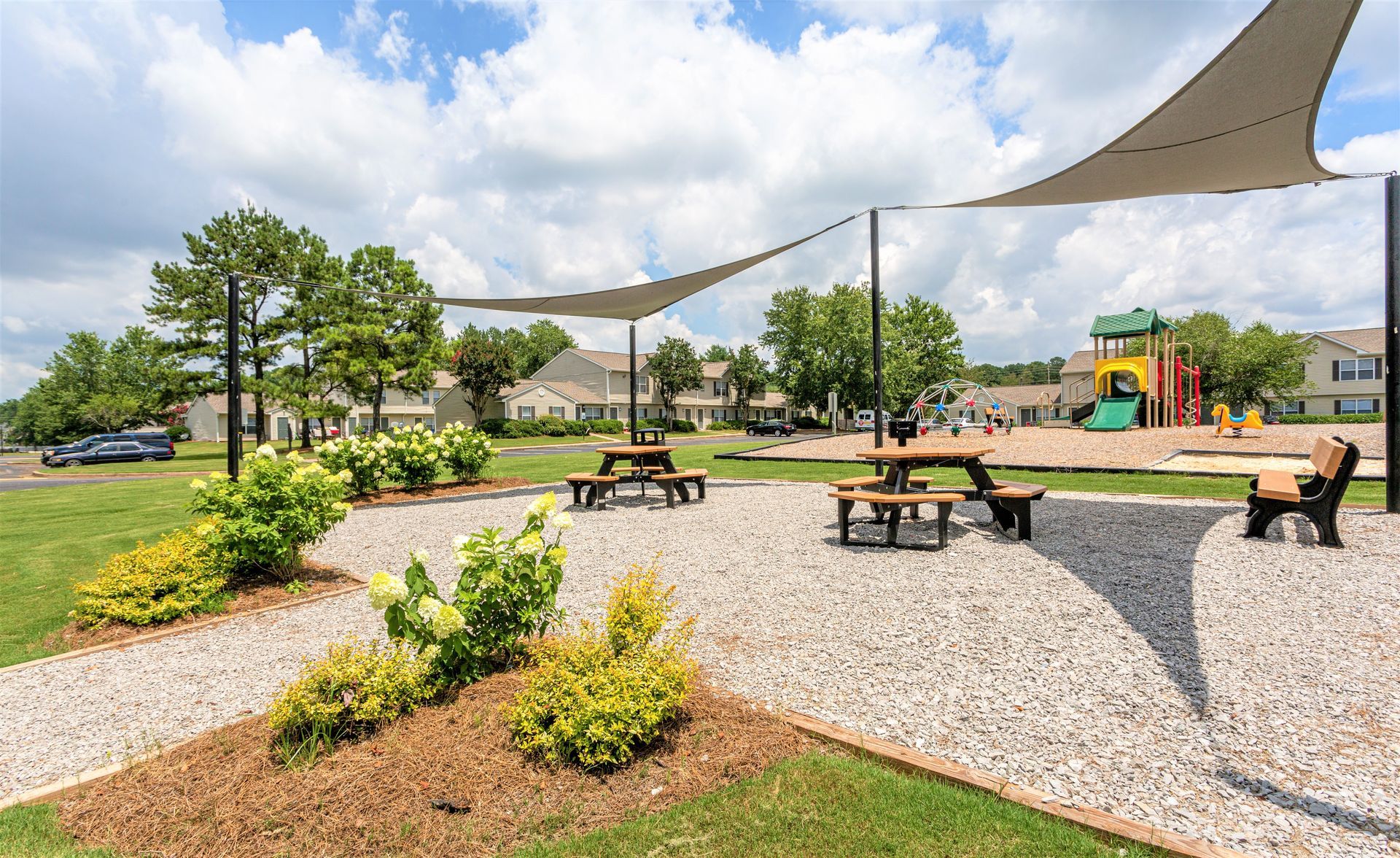 A park area with gravel ground, outdoor tables under shade sails, a small playground in the background, and green bushes.