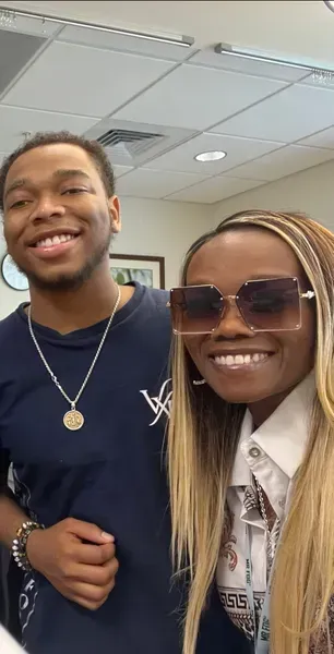 Two smiling people posing indoors, one in a navy shirt and one in sunglasses, under office ceiling lights