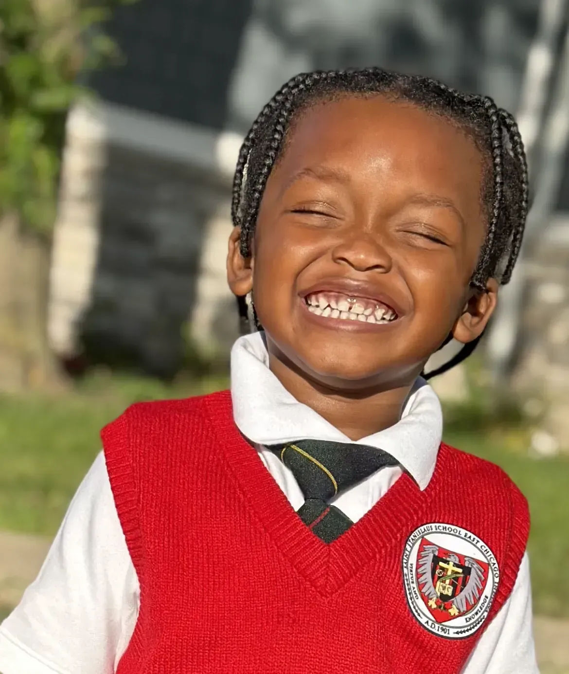 Smiling child in a red sweater vest and white shirt, standing outdoors in sunlight.