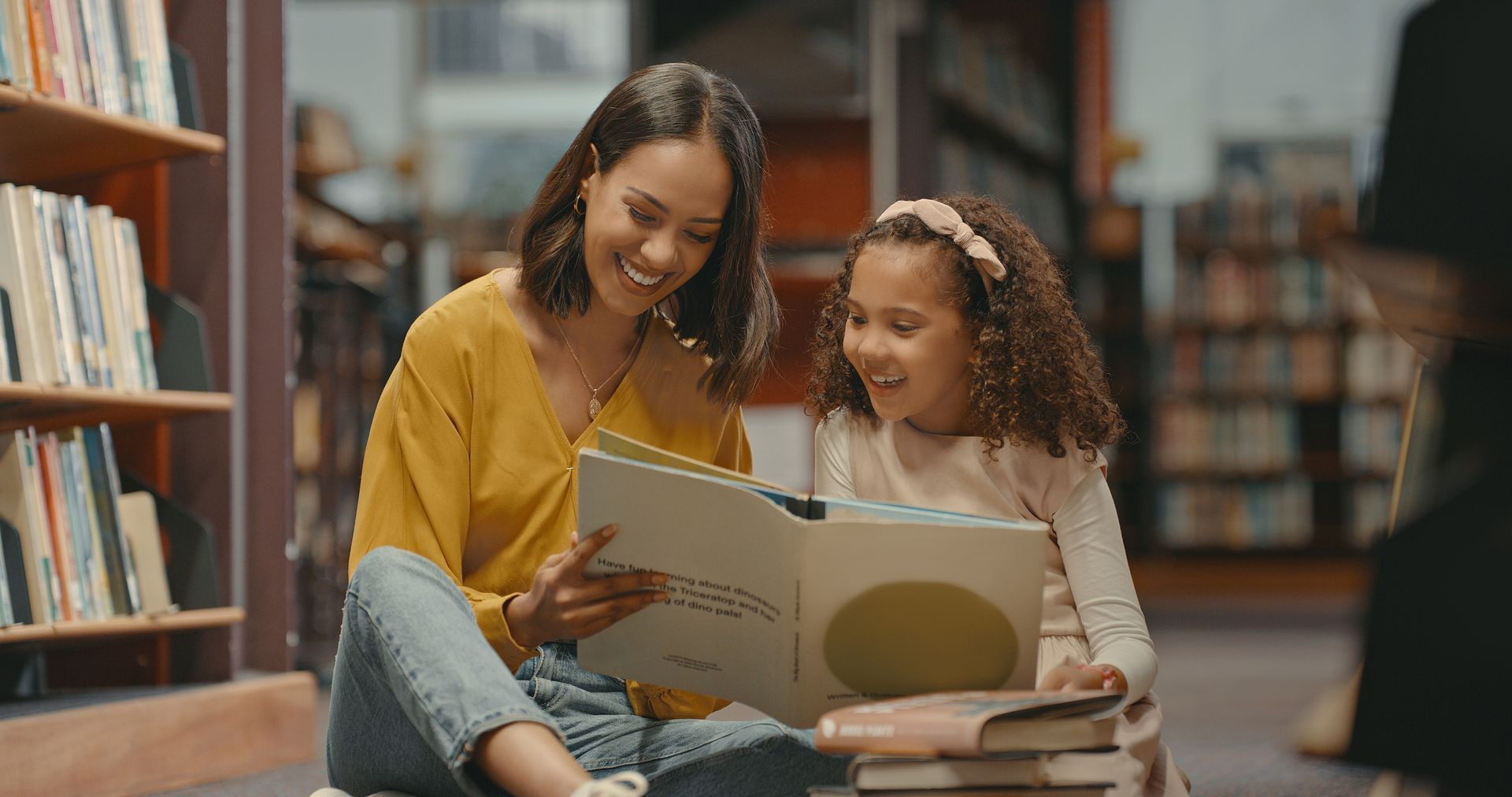 Adult and child reading a book together in a library, smiling on a cozy floor seating area.