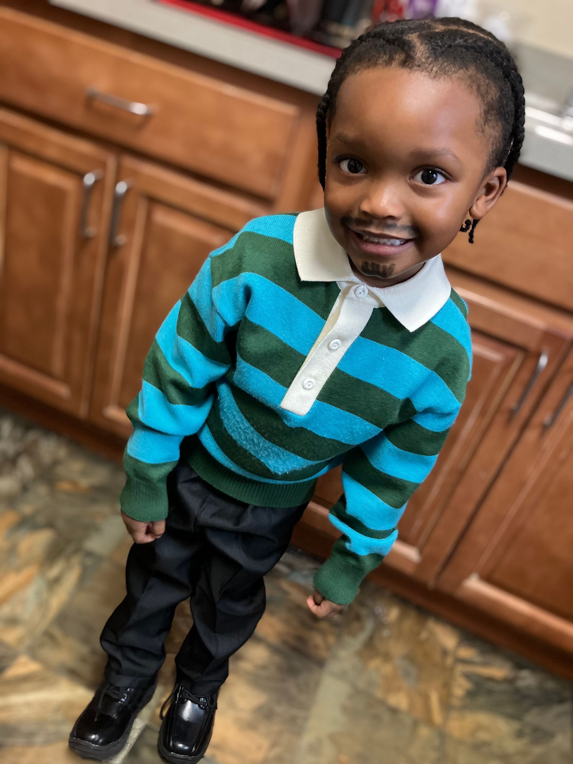 Child smiling in a turquoise striped sweater, black pants, and boots, standing in a kitchen.