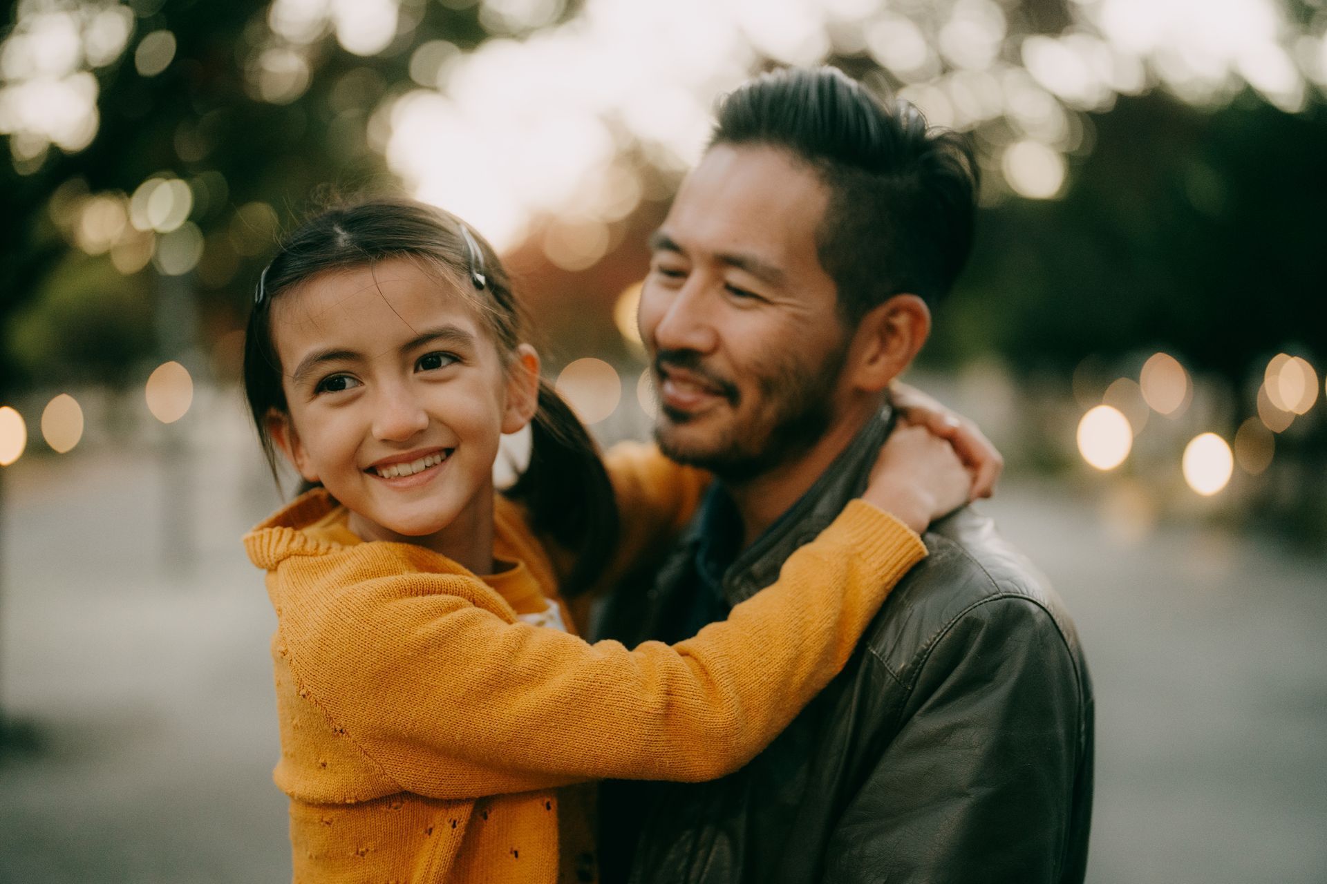 Smiling child in a yellow sweater hugging an adult outdoors at dusk with blurred lights in the background