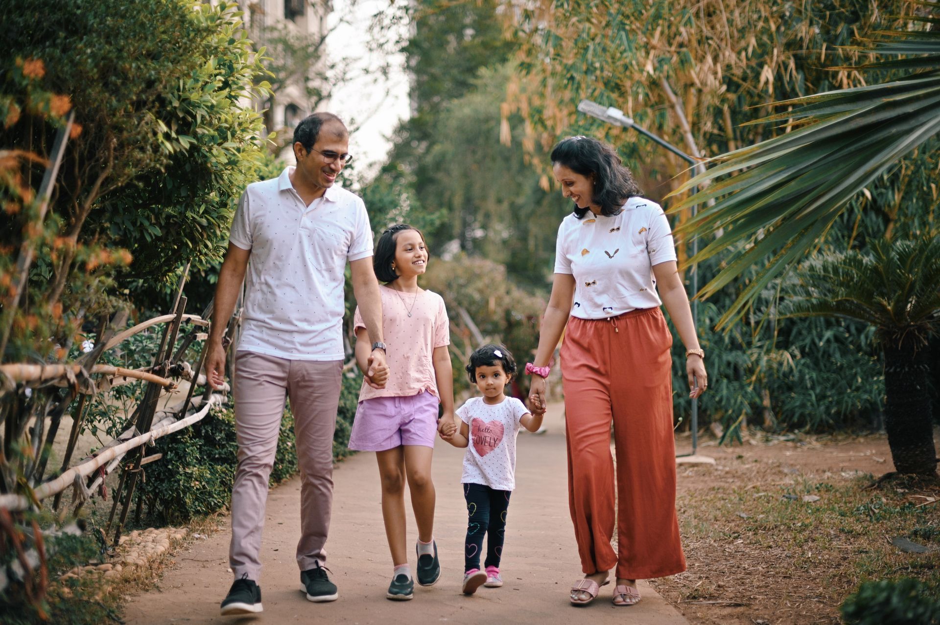Family walking together on a garden path, smiling and holding hands among trees.