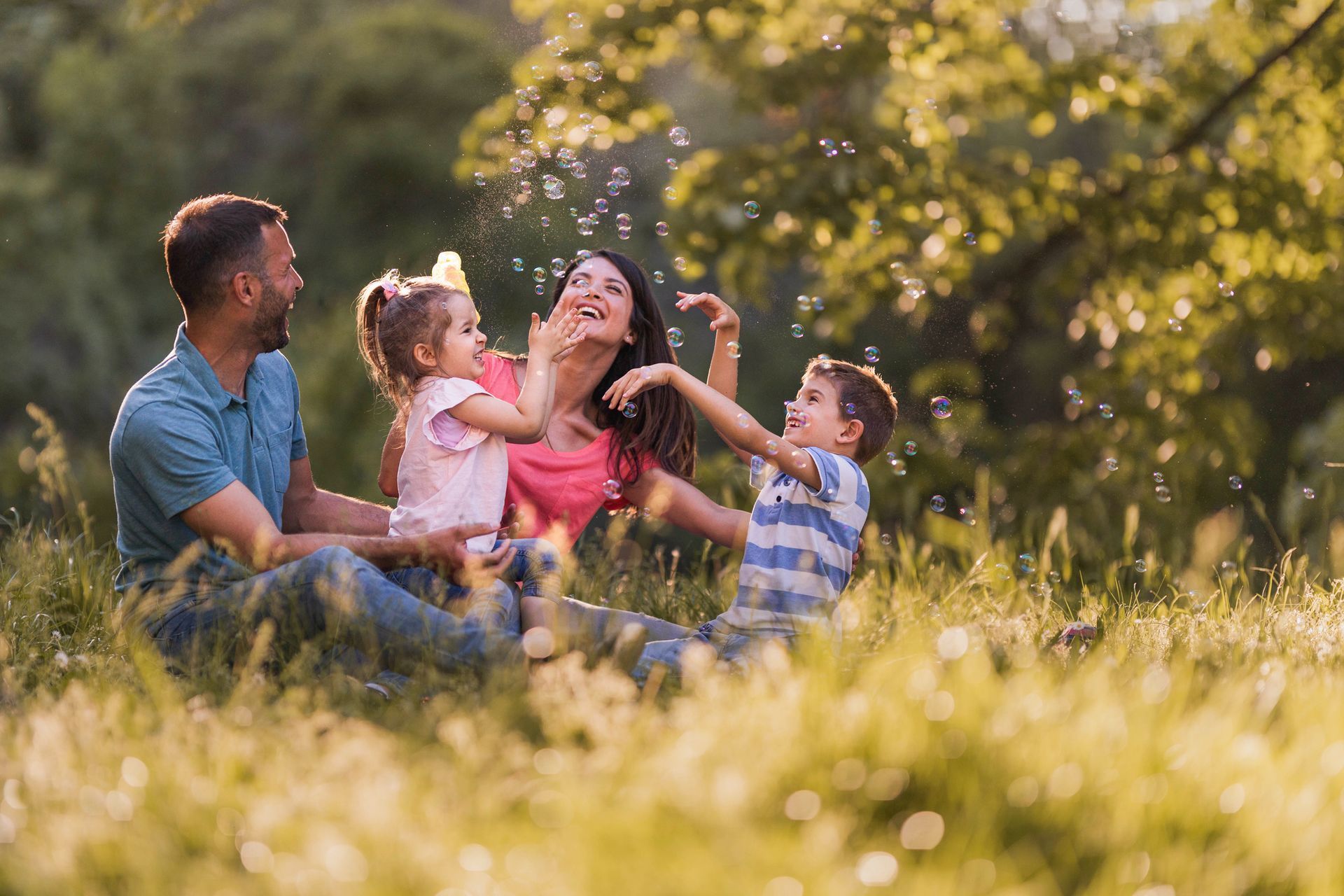 Family sitting in a sunlit meadow, laughing and playing together with a young child.