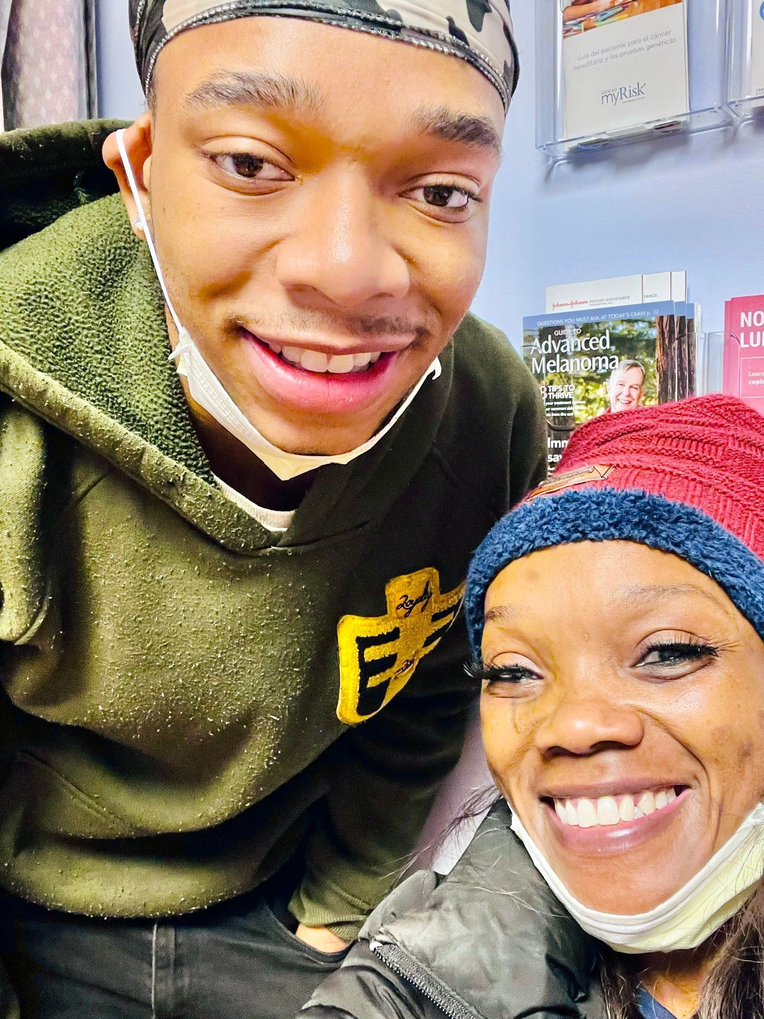 Two smiling people indoors, one in a green sweater and one in a red beanie with a mask under her chin.