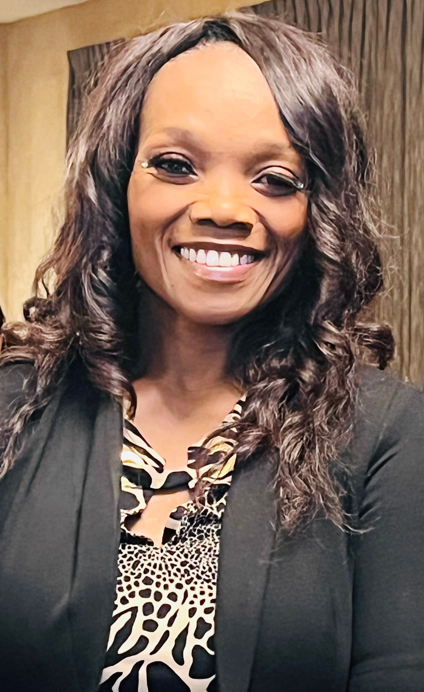 Smiling woman in a black blazer and patterned blouse, standing indoors against a wood-paneled background