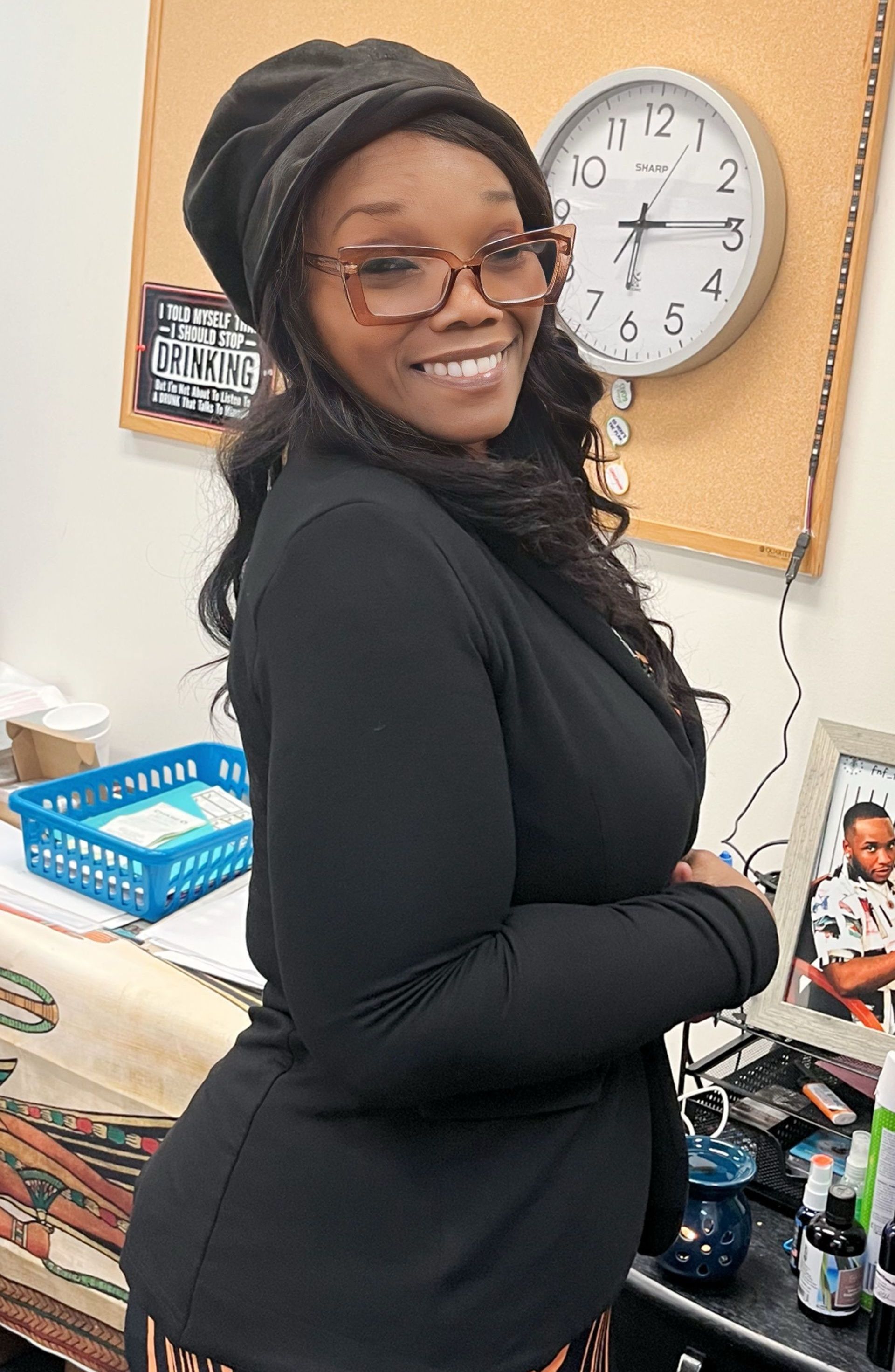 Smiling person in black clothing and glasses standing in an office beside a desk and wall clock