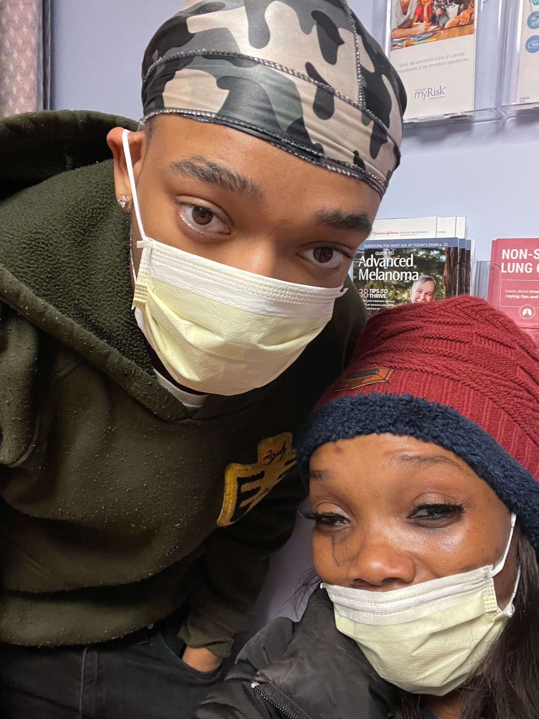 Two masked people leaning together indoors, one in a camo headscarf and green jacket, one in a red knit hat.