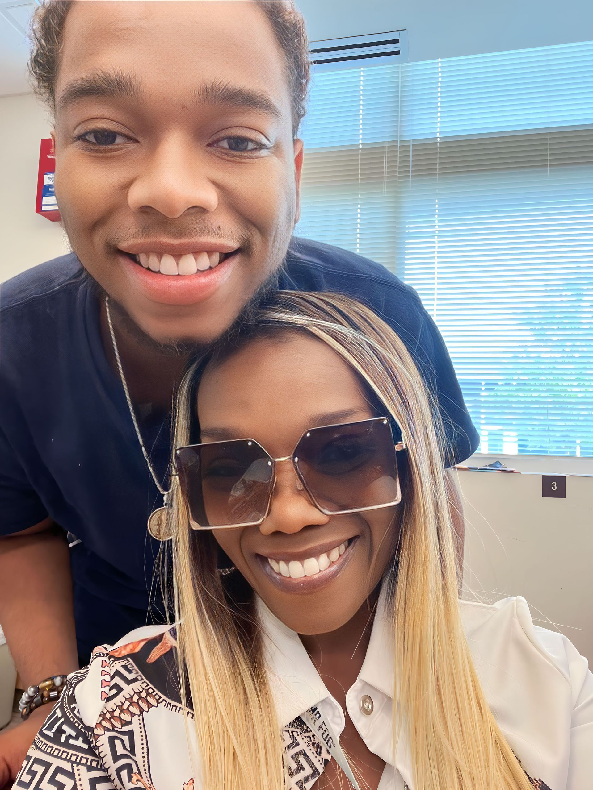 Two smiling people posing indoors, one wearing sunglasses, in front of window blinds.