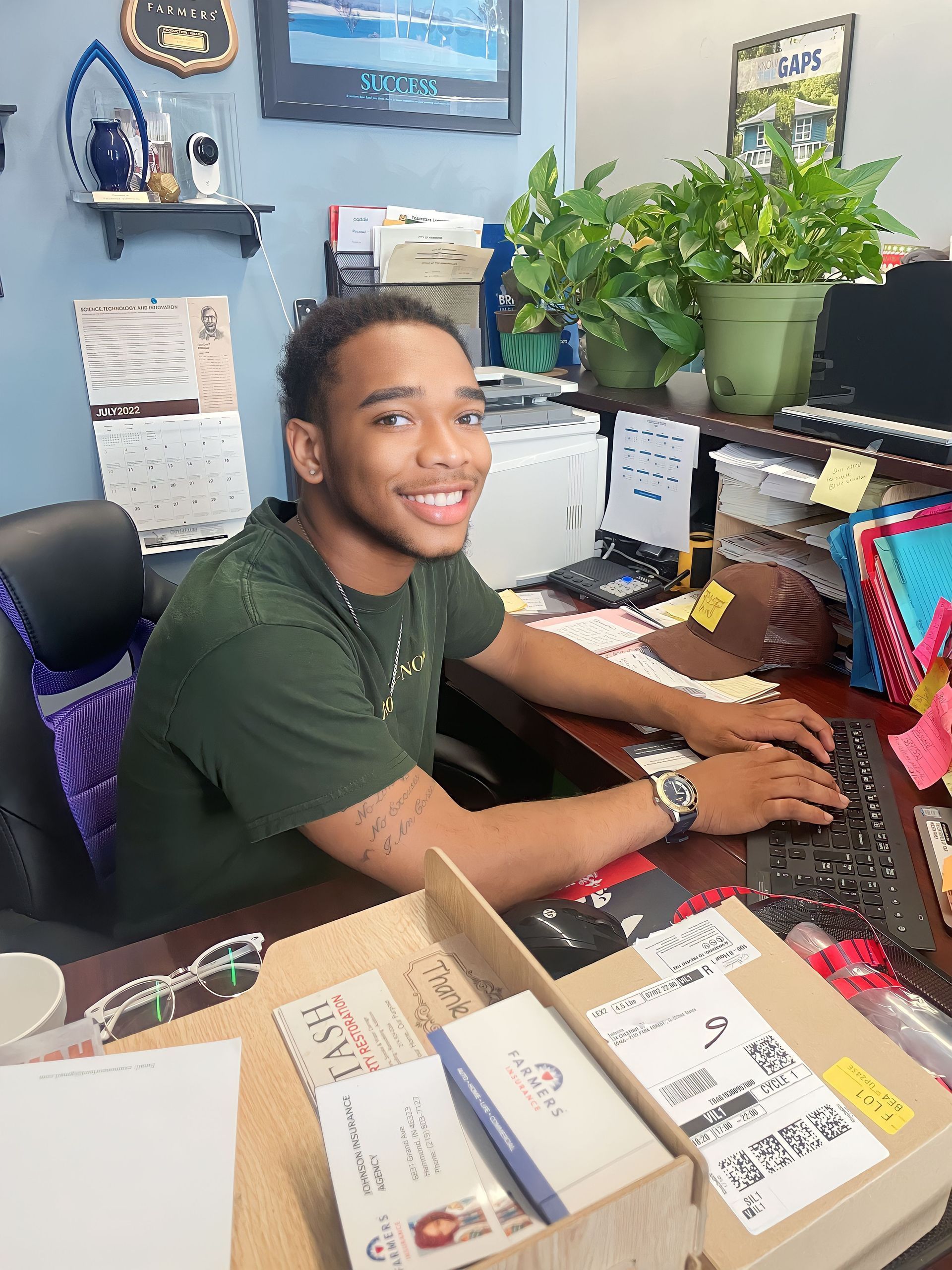 Smiling person at a desk with computer, keyboard, paperwork, and potted plants in a home office