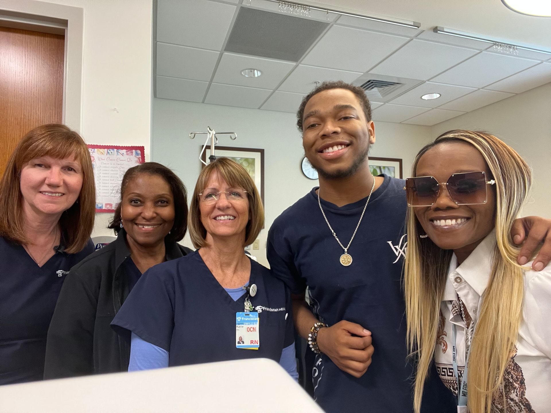 Five smiling coworkers posing together in an office hallway, two wearing badges and one in sunglasses.