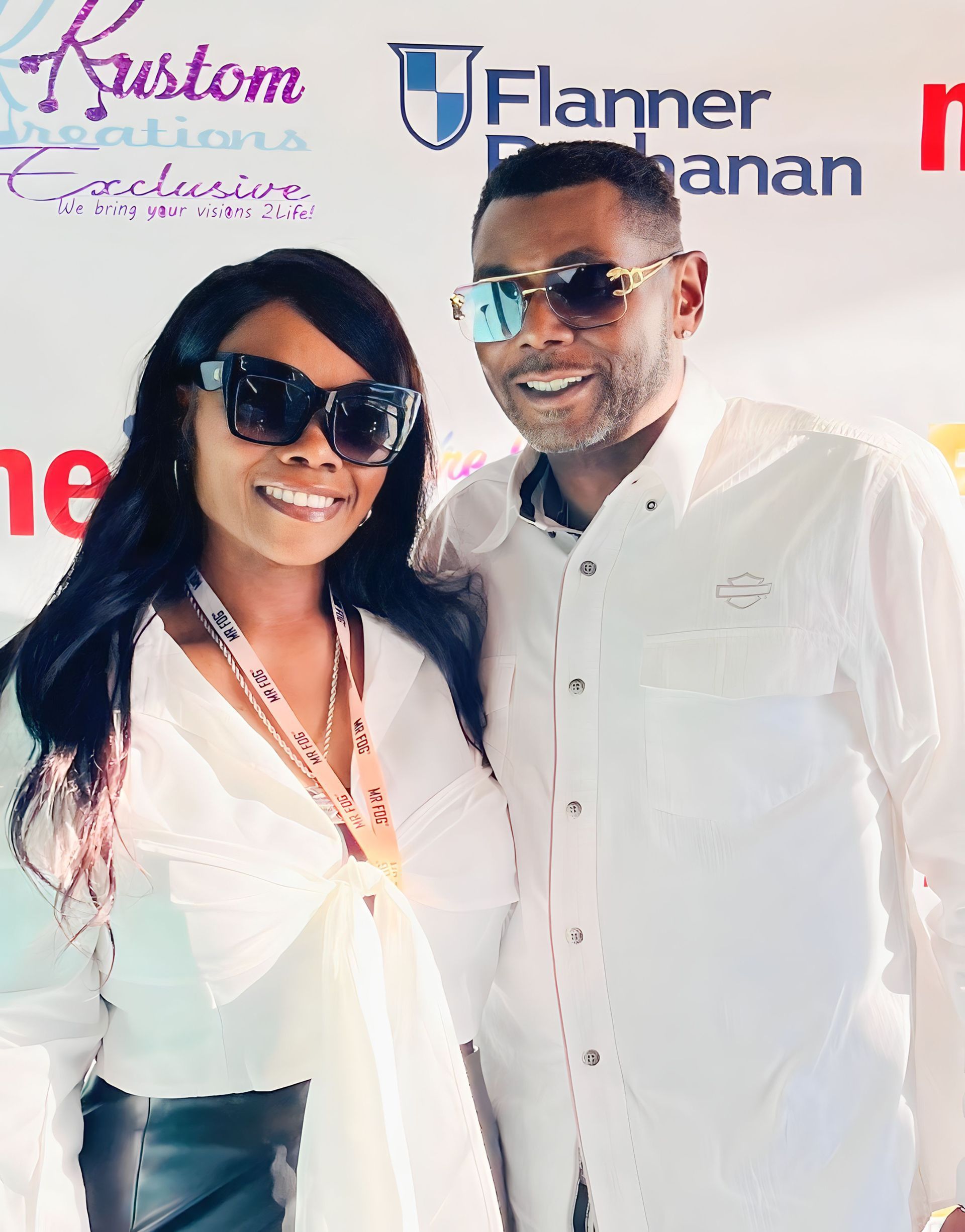Two people smiling in white outfits and sunglasses at a branded event backdrop.