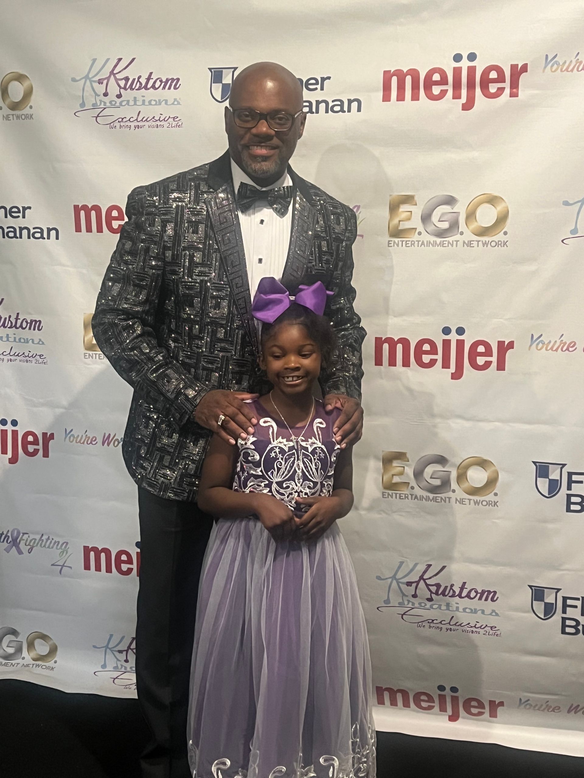 Man and young girl posing together on a red carpet in formal attire, smiling before a sponsor backdrop