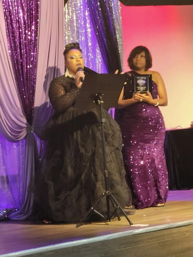 Two women in purple gowns on stage, one speaking at a podium and the other holding an award.
