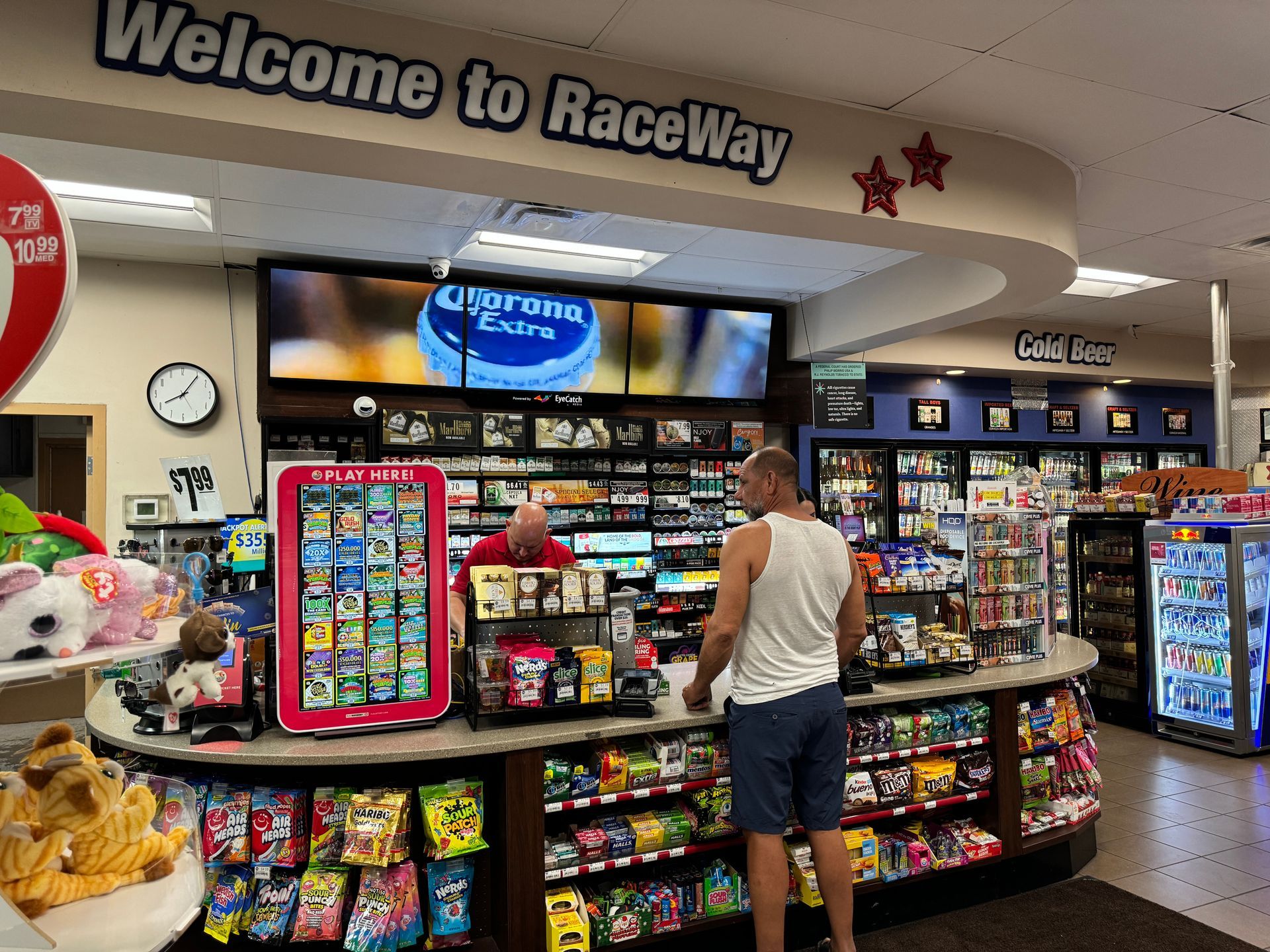 Convenience store checkout with candy displays and a customer watching a Corona ad on digital  in-store screens.