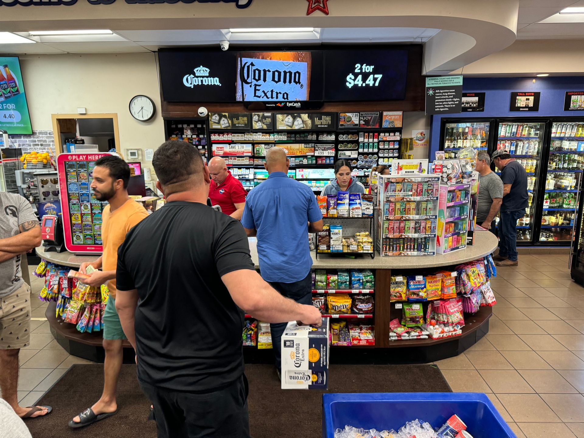 People waiting in line at a convenience store, watching a Corona ad on digital screens.