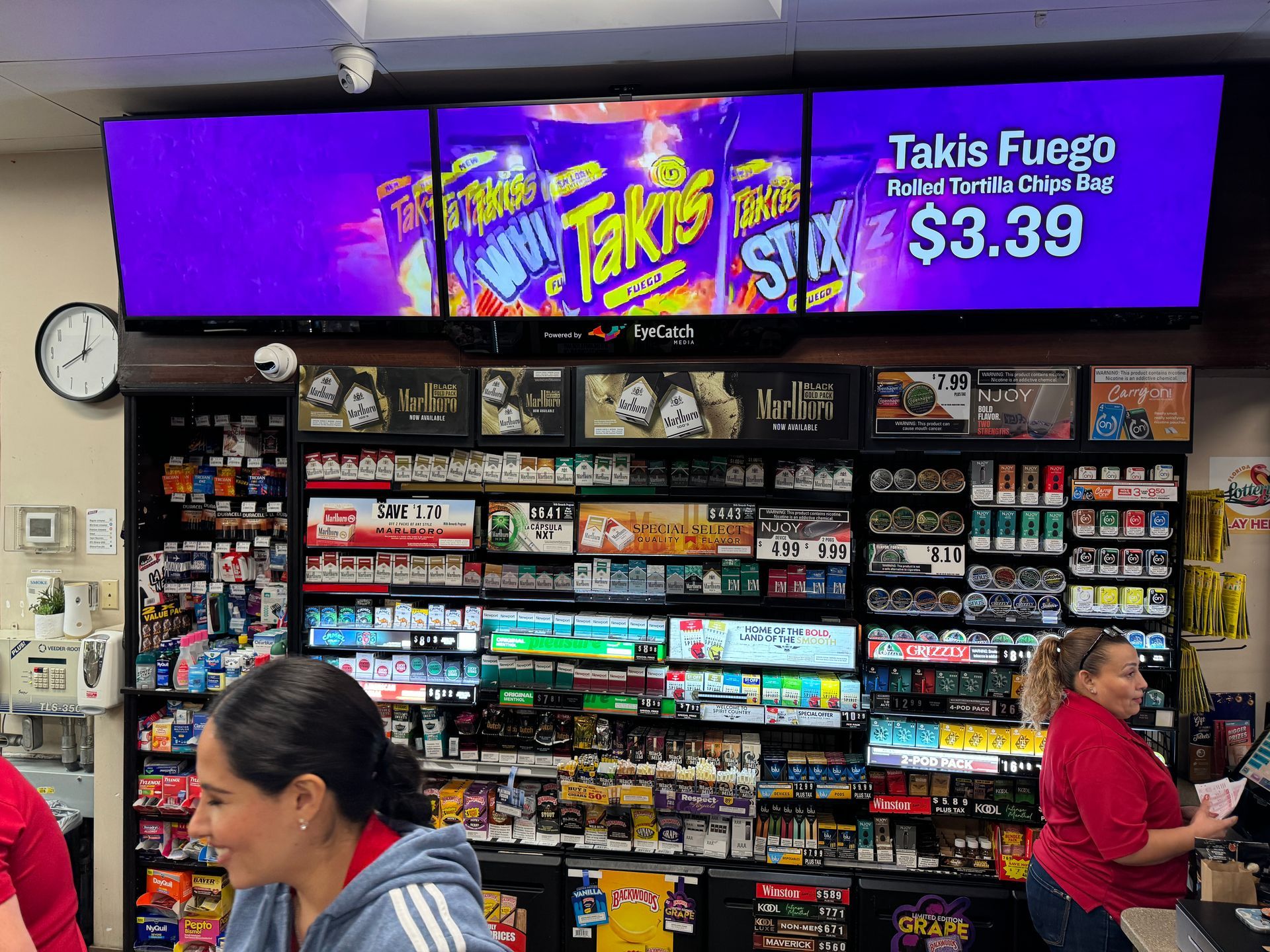 Convenience store snack aisle with Takis Fireo sign and shoppers in red shirts