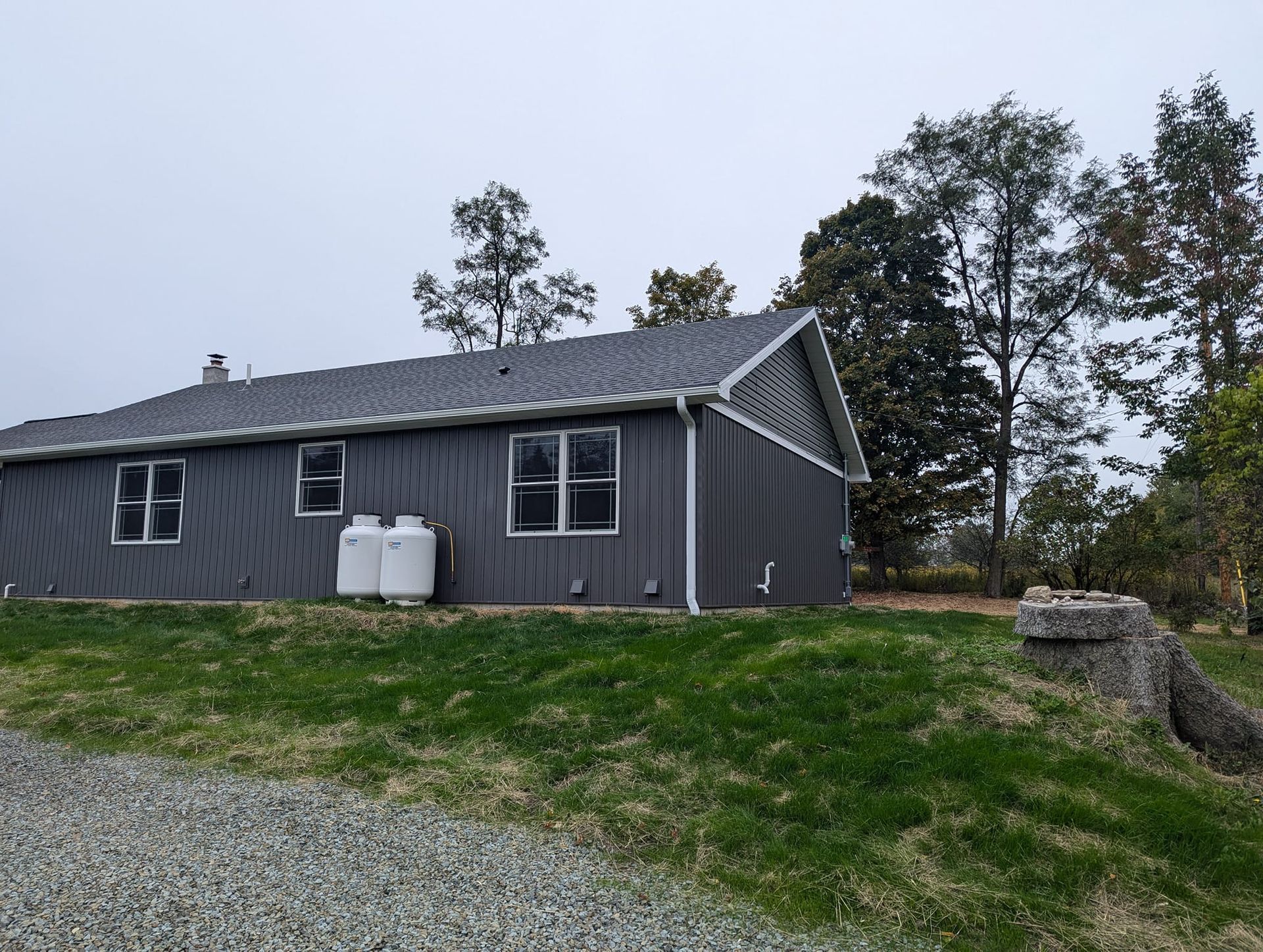 Grey house with propane tanks on a grassy hill; trees in the background under an overcast sky.