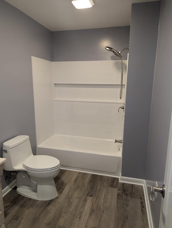 Bathroom with gray walls, white toilet, bathtub, and shower fixture. Gray wood-look flooring.