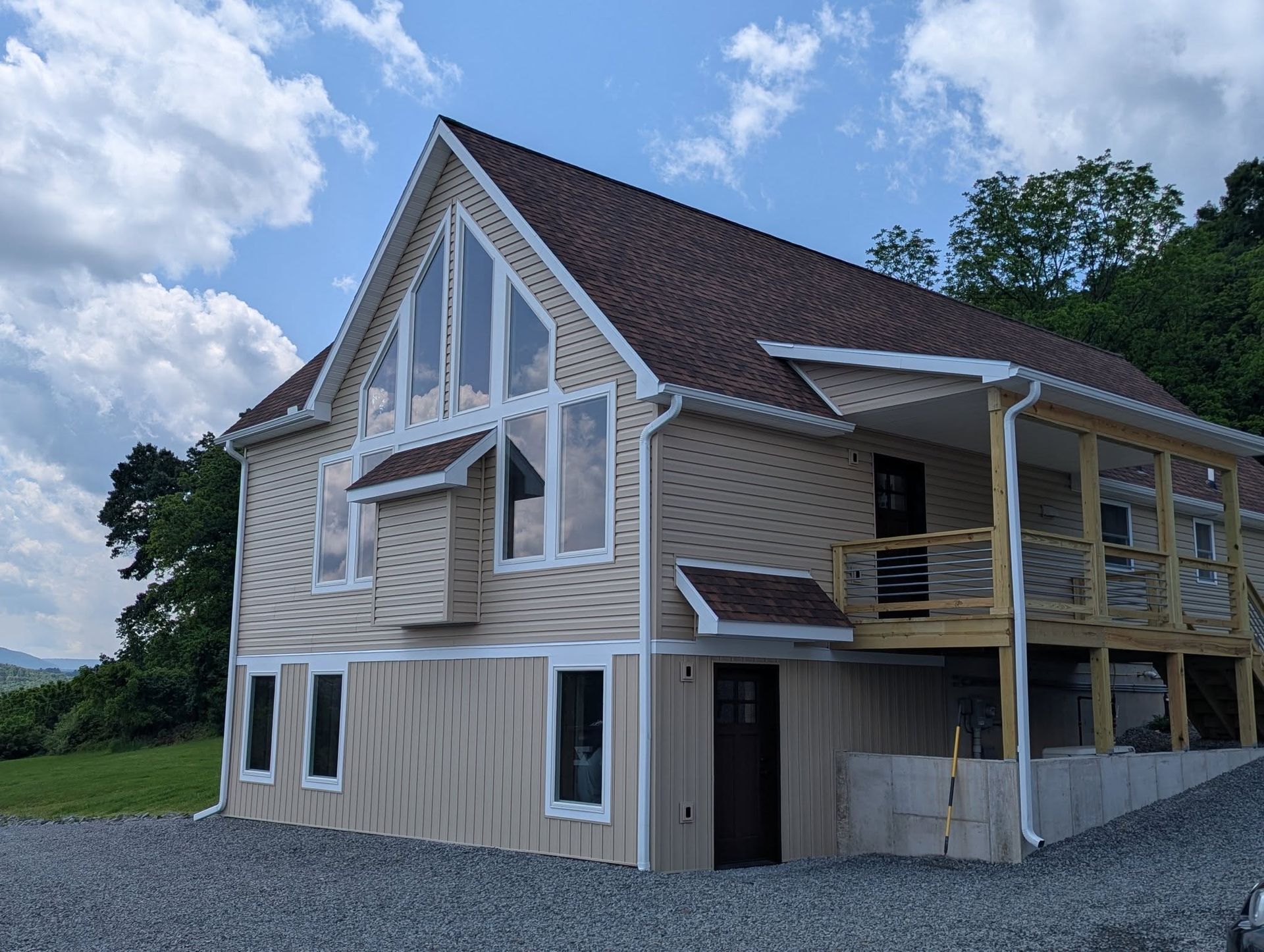 Two-story house with beige siding, brown roof, large windows, and a wooden deck under a cloudy sky.