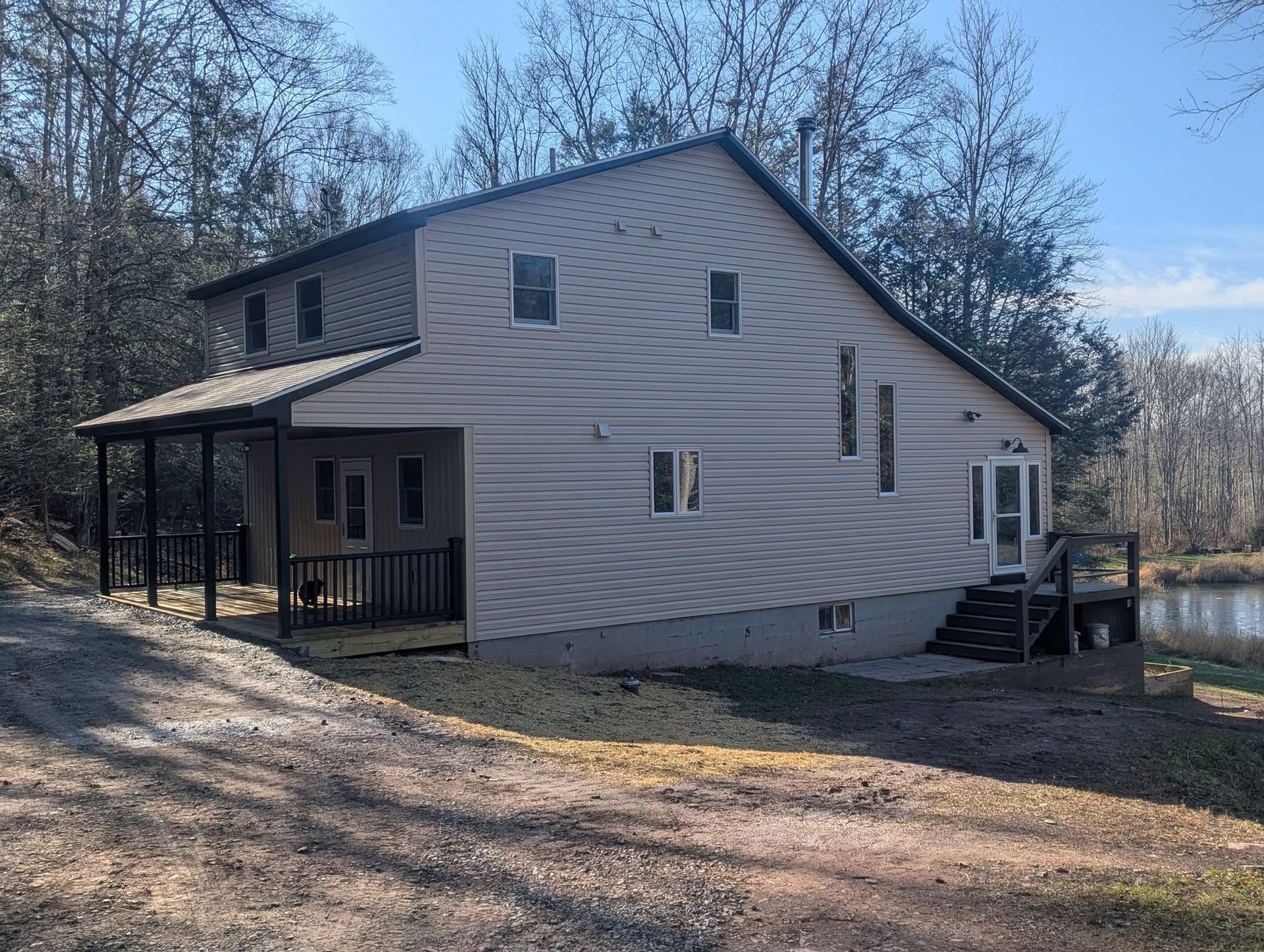 Two-story house with gray siding, porch, and metal roof near trees and water.