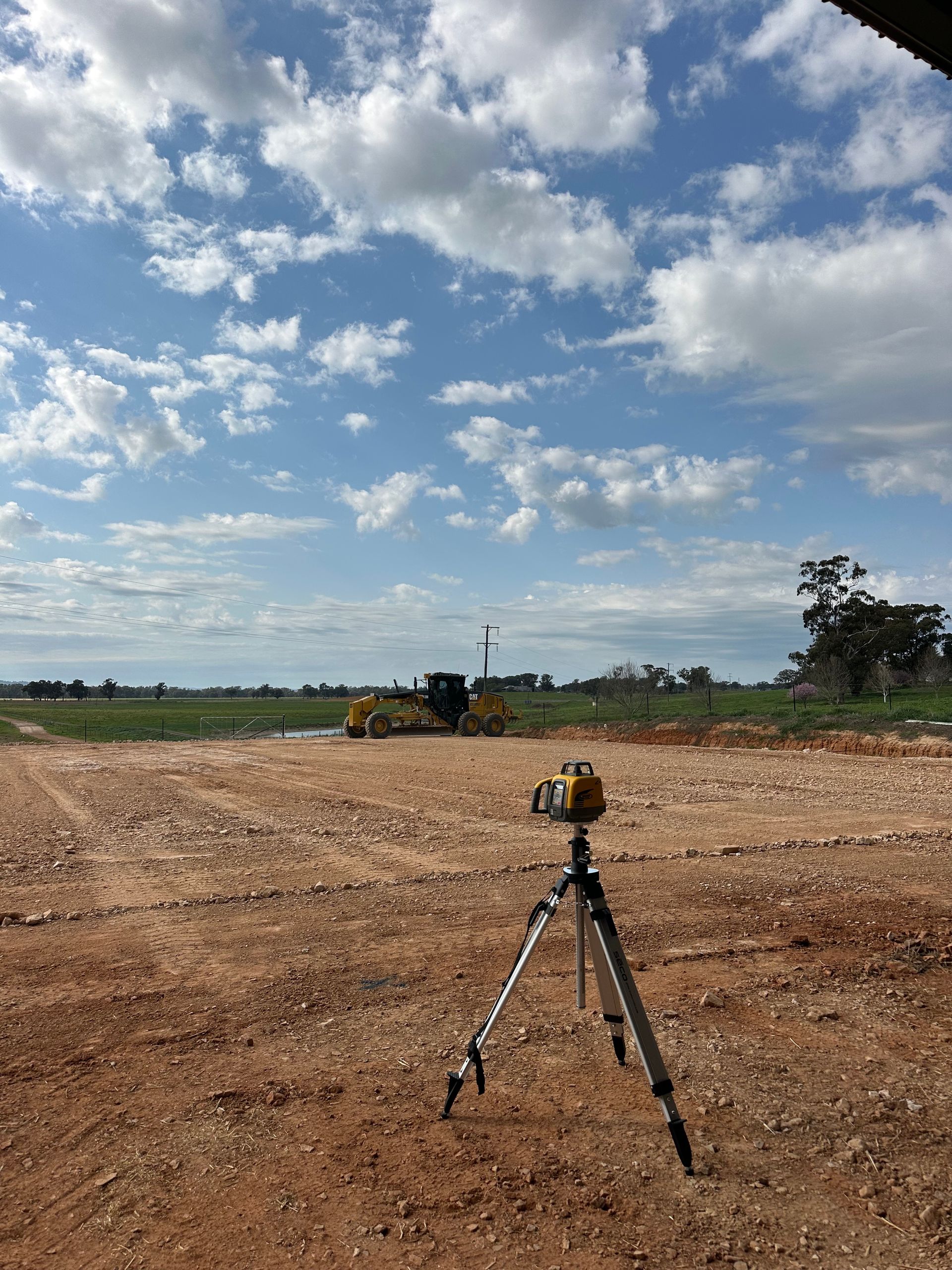 Laser level on tripod at civil construction site with grader ready, precision earthworks and site leveling in progress.