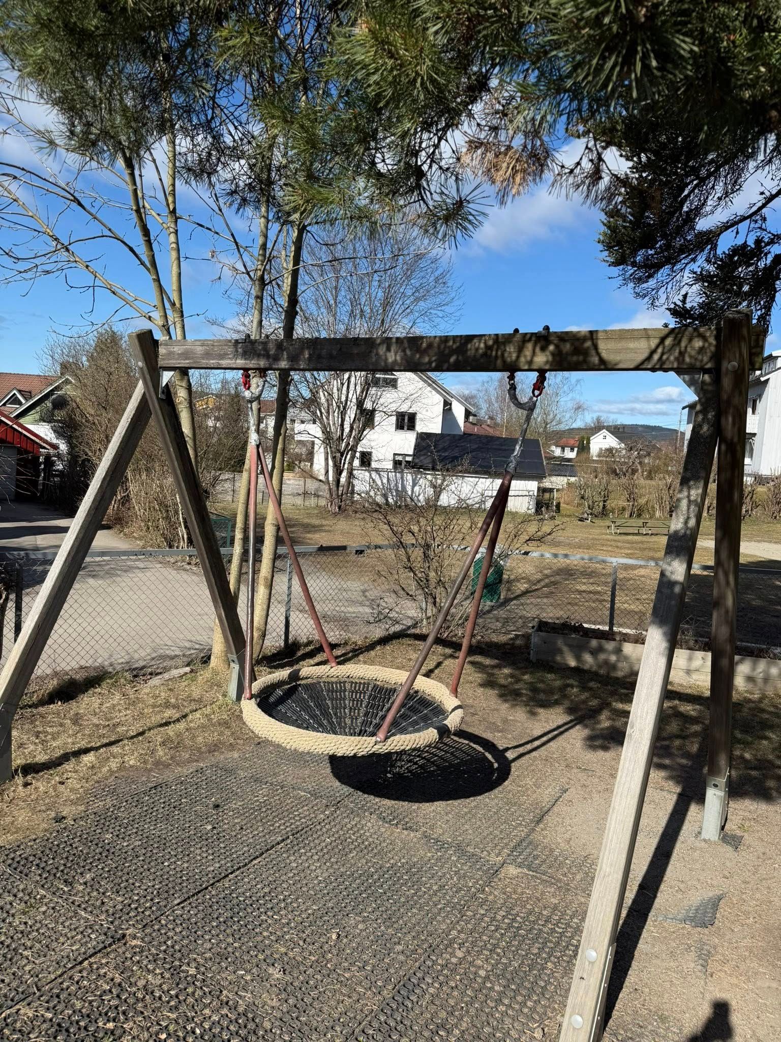 A wooden swing set with a round nest swing on a gravel playground under a clear blue sky.