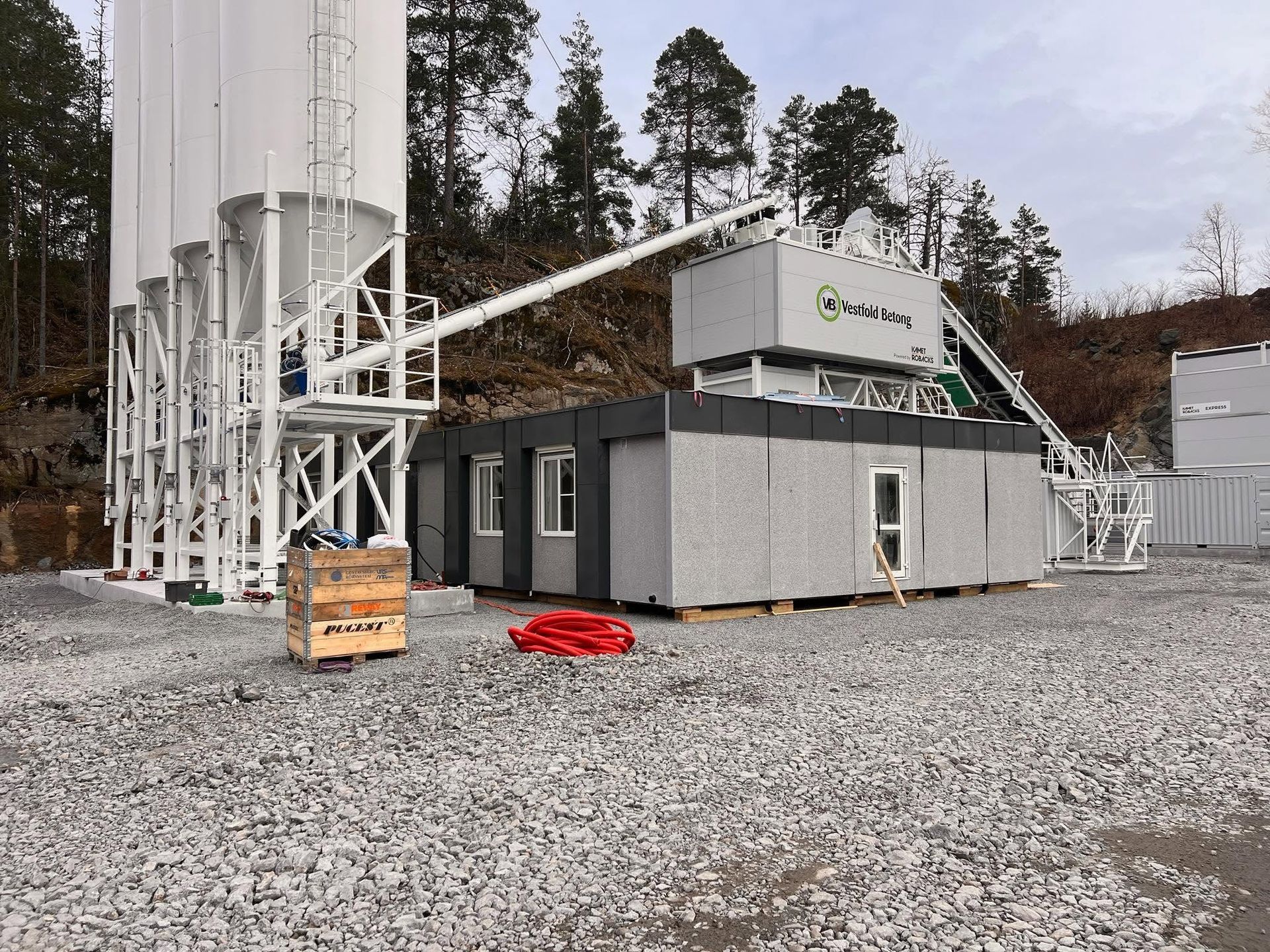 Small industrial building beside silos on a gravel lot, with trees and a hillside in the background.