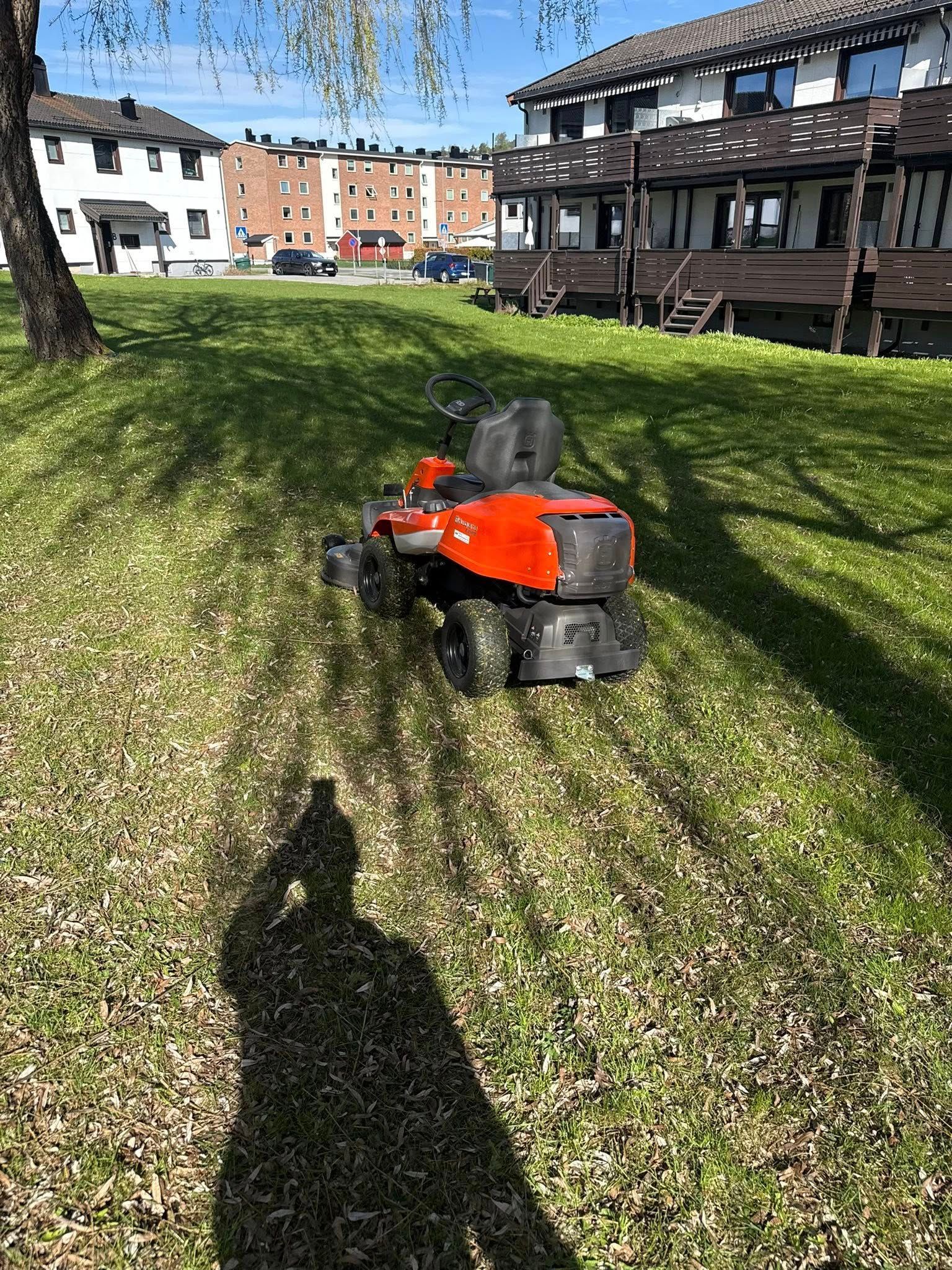 Orange lawn mower on a grassy slope in a sunny park, with buildings in the background.