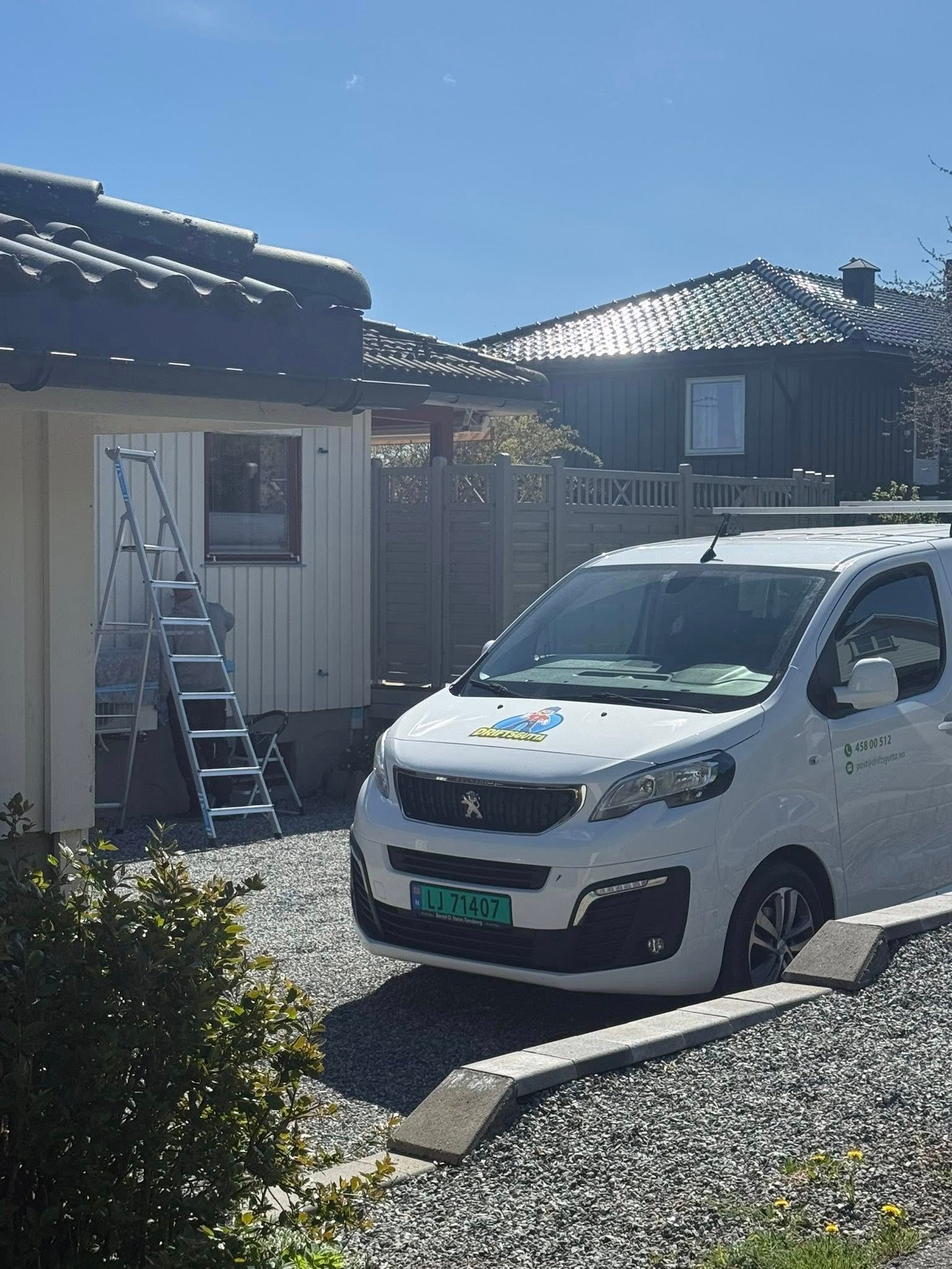 Hvit varebil parkert i en solrik grusinnkjørsel ved siden av et lite hus og en stige under en blå himmel