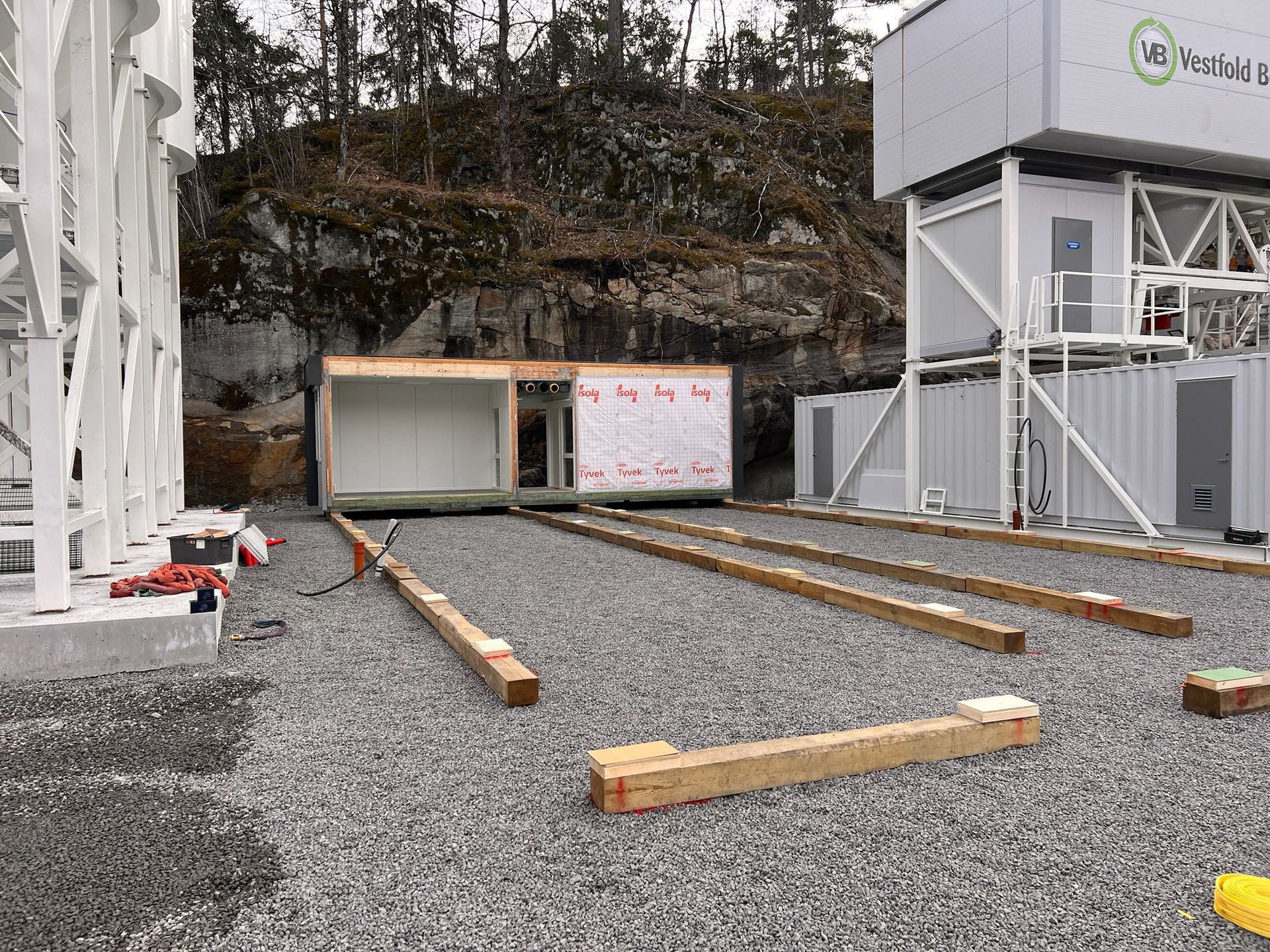 Construction site with gravel ground, wooden beams, and a small building beside a large white structure