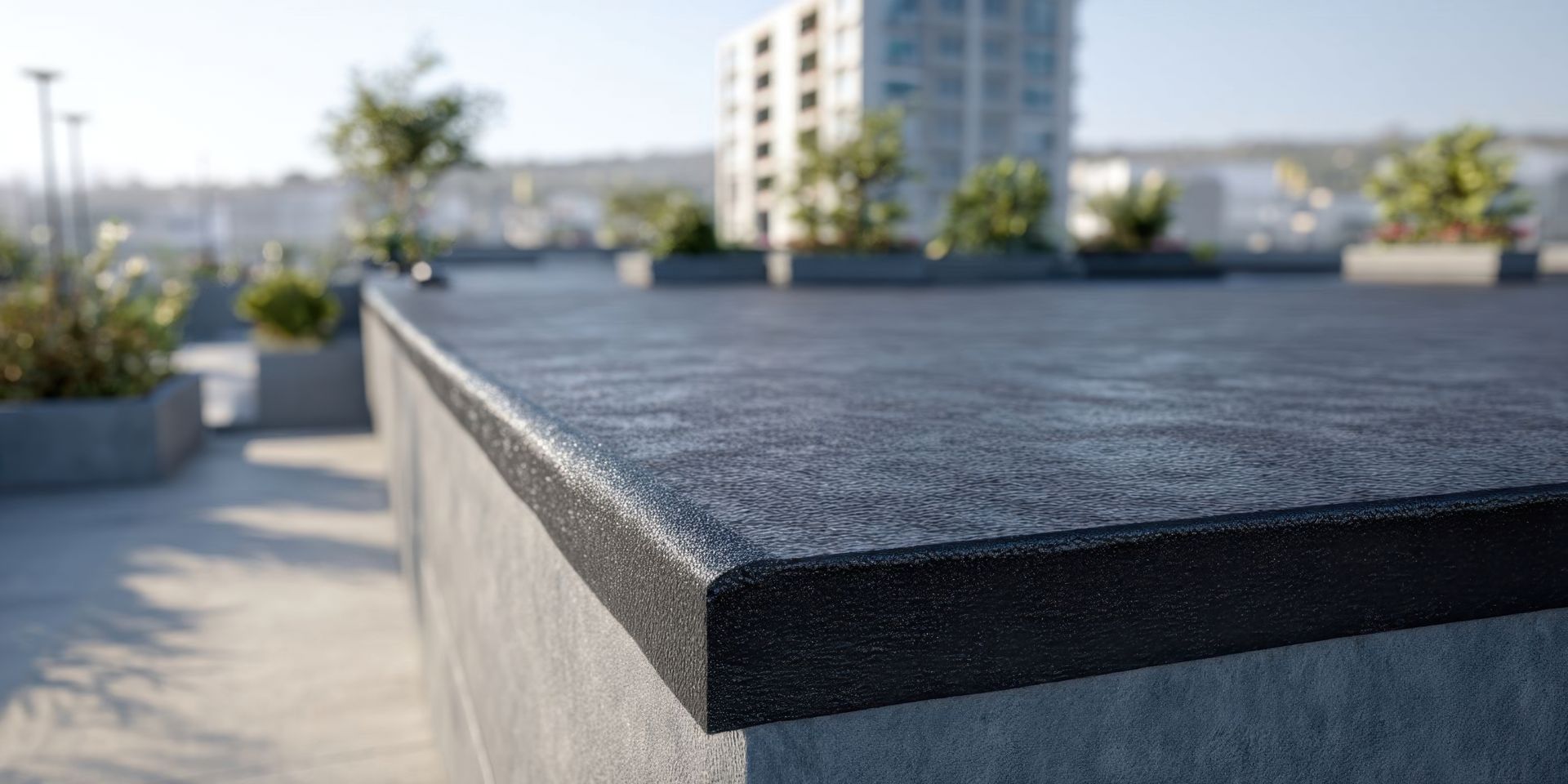 Gros plan sur un banc ou un rebord noir en extérieur, sur une terrasse ensoleillée sur un toit, avec des immeubles de la ville flous en arrière-plan.