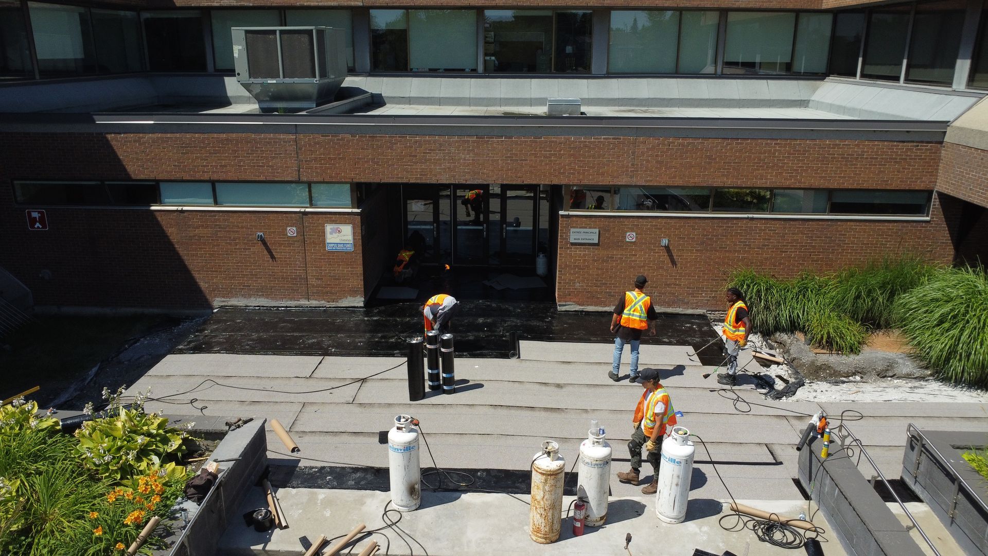Des ouvriers en gilets orange travaillent sur le patio d'entrée d'un bâtiment en briques, avec des outils et des barrières au premier plan.