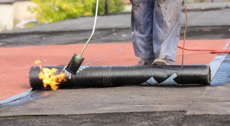 Une personne utilise une torche pour chauffer un rouleau de toiture noire sur un toit, les flammes se trouvant à l'extrémité gauche.