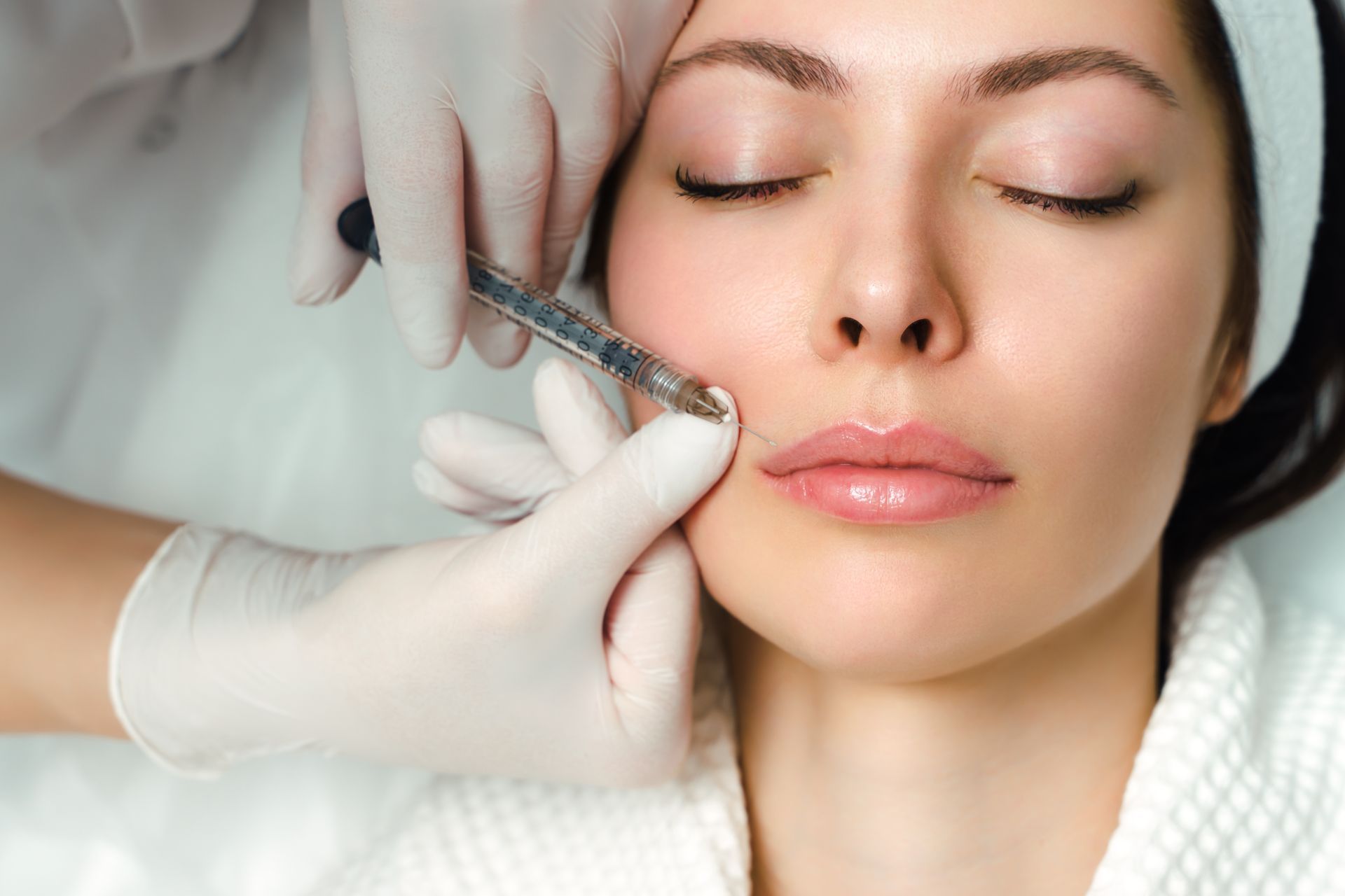 Person receiving lip filler injection. Hands wearing gloves hold syringe near mouth.