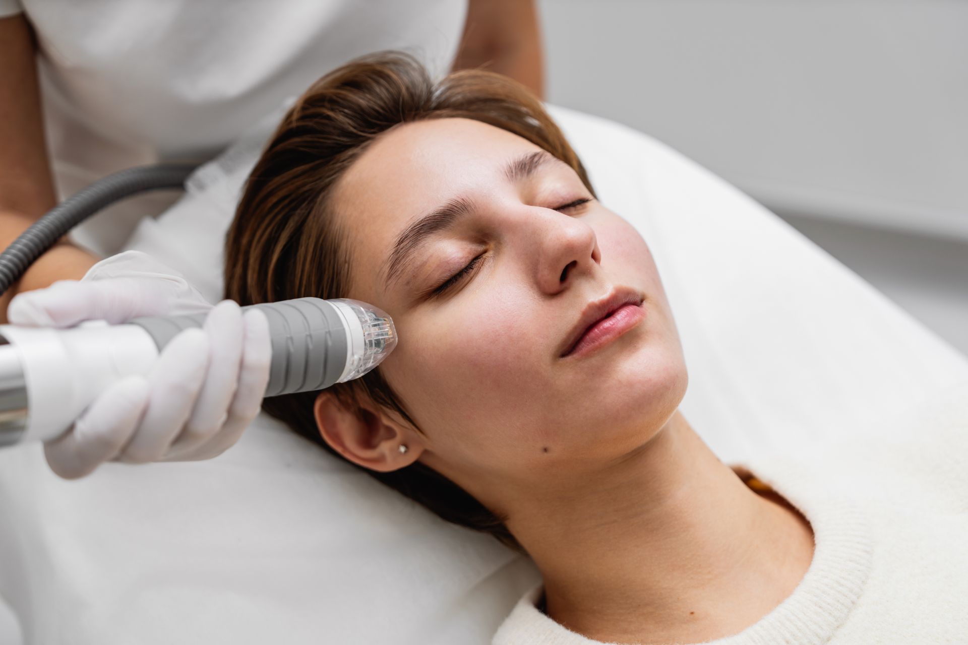 Woman receiving facial treatment in a clinic; a medical device is held to her face.