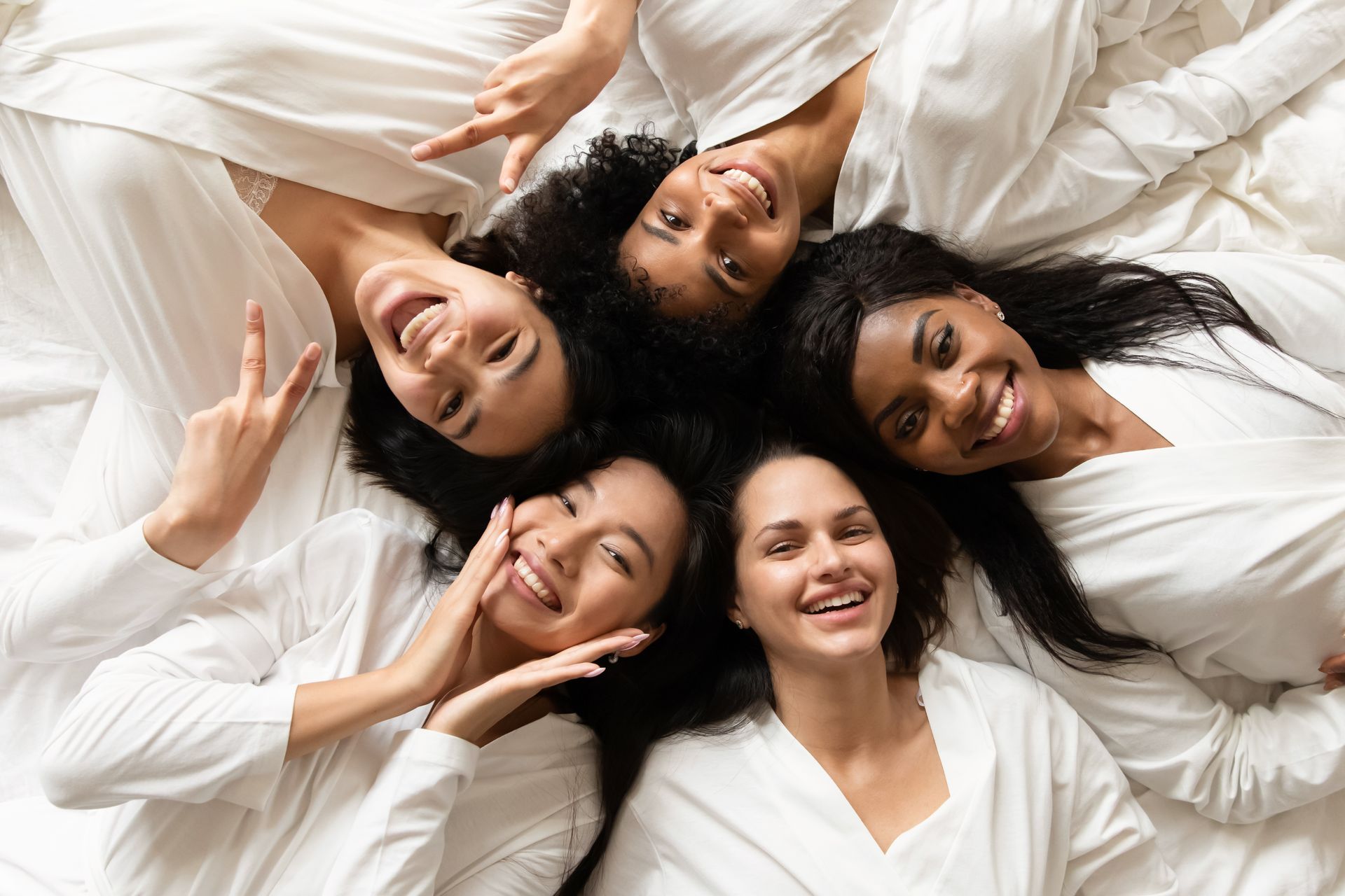 Five people lying in a circle on white bedding, smiling and resting their heads together