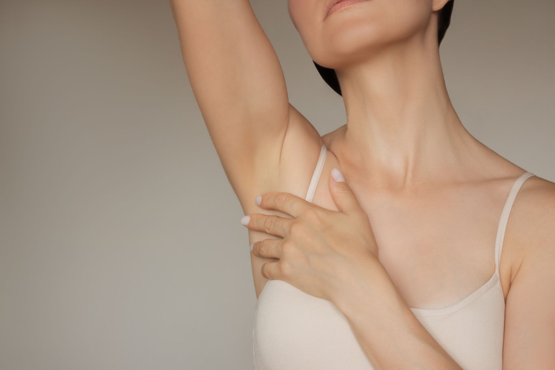 Woman raising arm, hand touching underarm, wearing a tank top, against a neutral background.